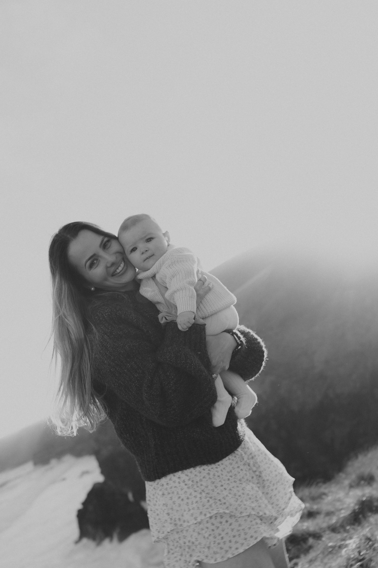 Black and white photo of a smiling woman holding a baby outdoors with misty mountain backdrop.