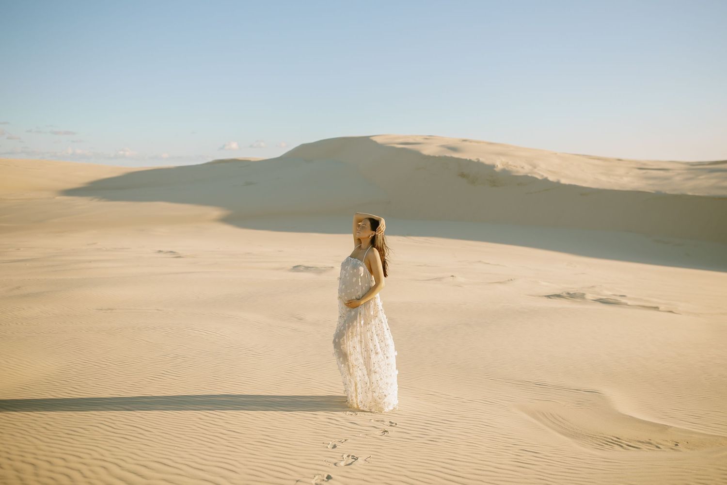 Figure in a flowing white dress stands on vast sand dunes under clear blue sky in desert landscape.
