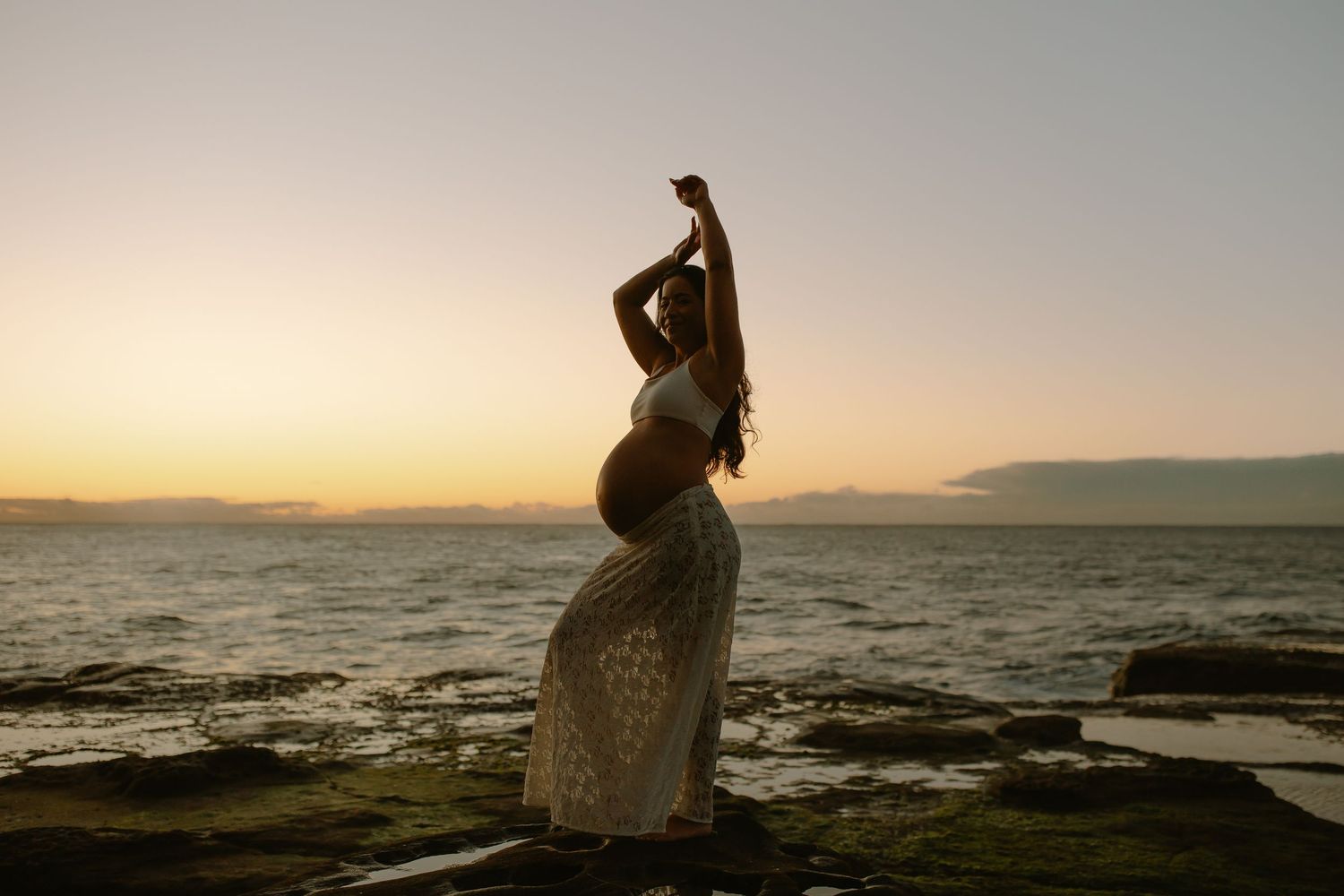 A silhouetted pregnant woman in a lace dress poses gracefully against a sunset ocean backdrop.