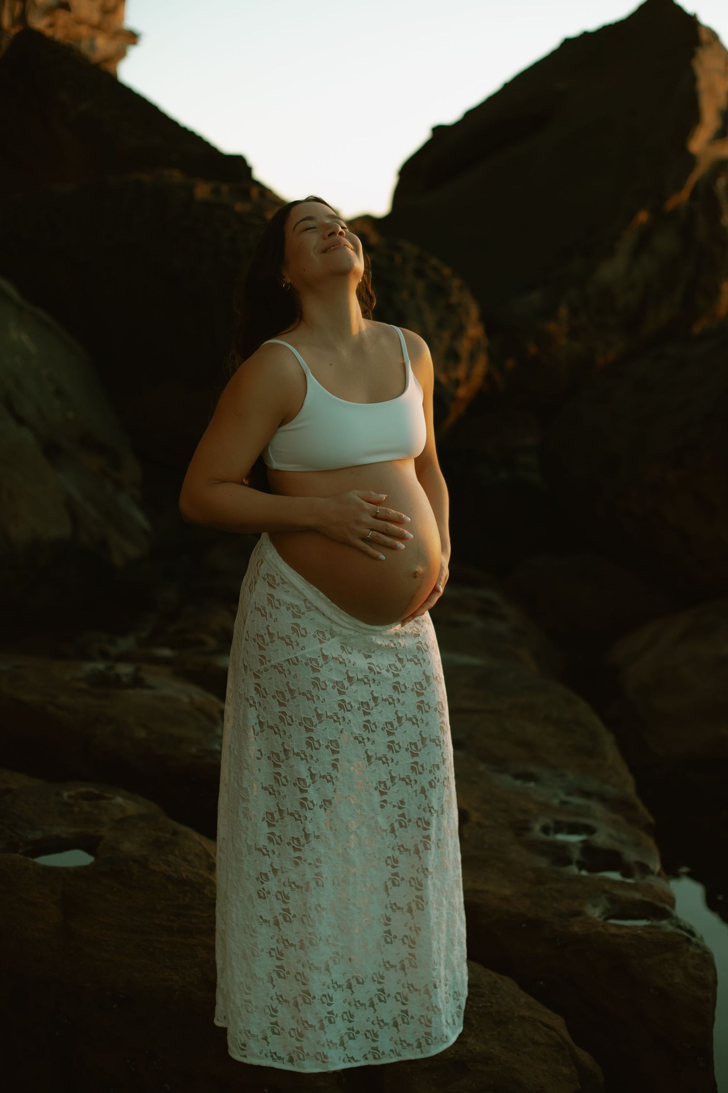 Silhouetted pregnant woman in white lace dress poses against rocky cliffs at sunset.