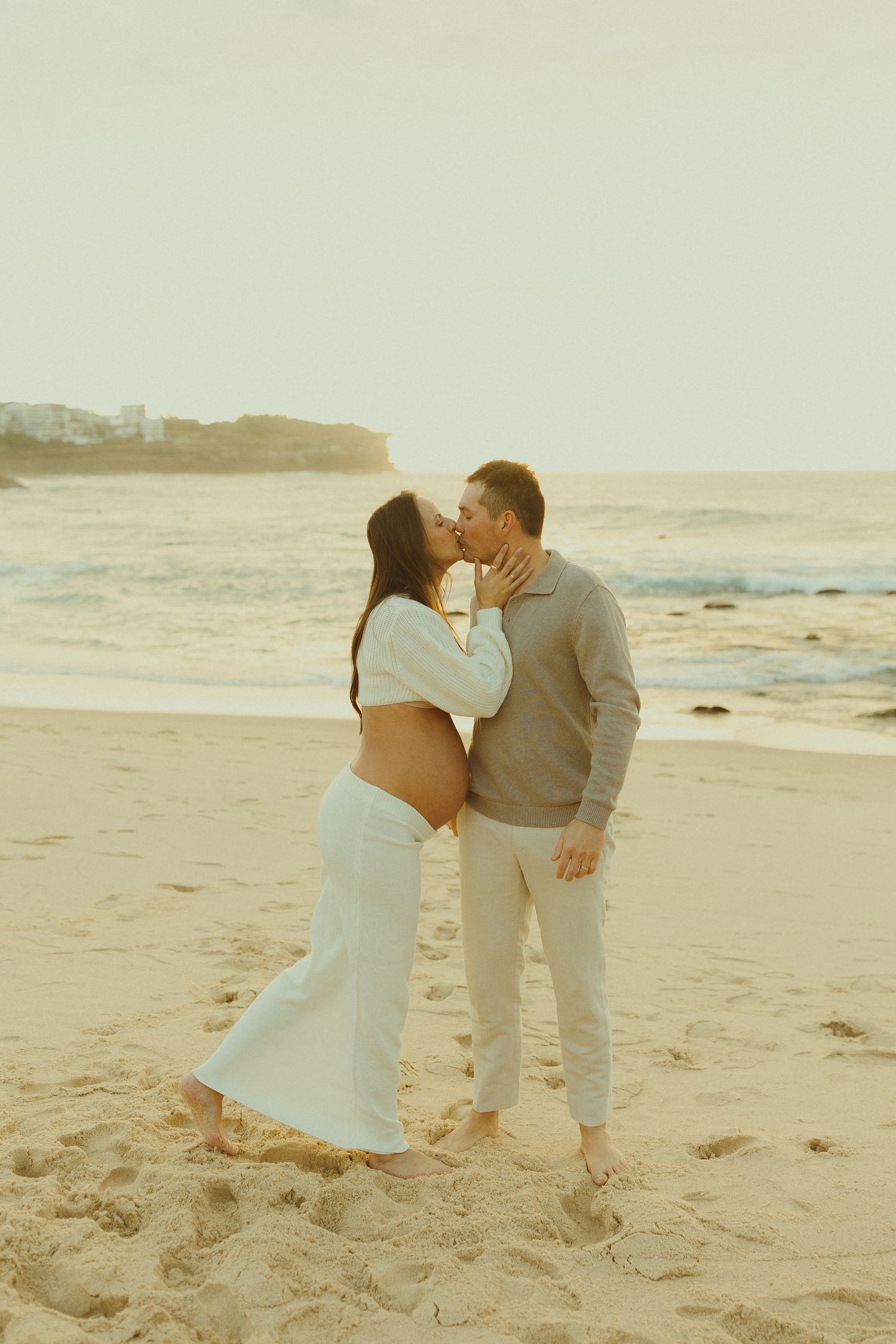A couple sharing a romantic kiss on a sandy beach at sunset wearing neutral colored outfits.