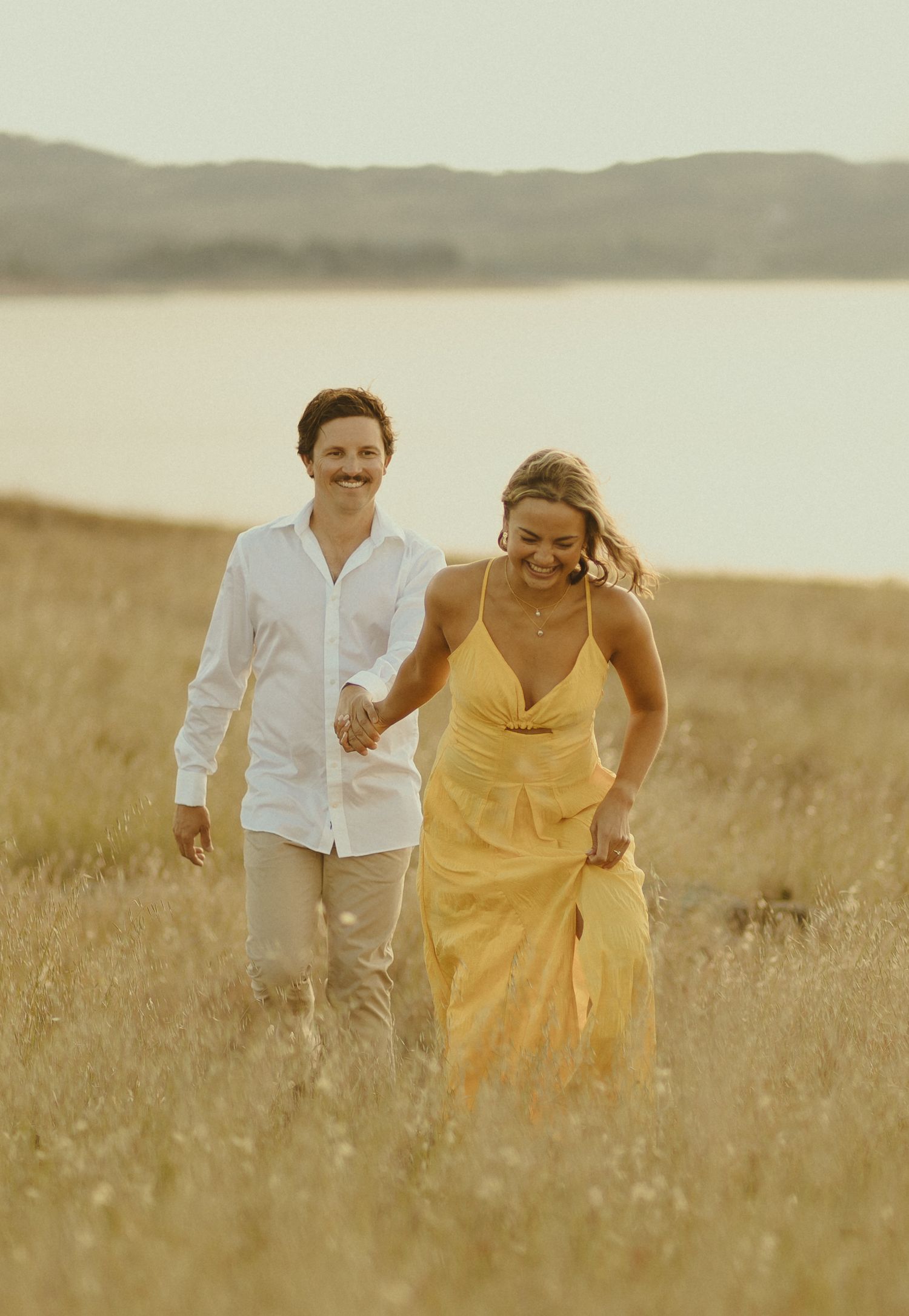 A couple walks through golden grass near a misty lake at sunset, with one wearing yellow and one in white.