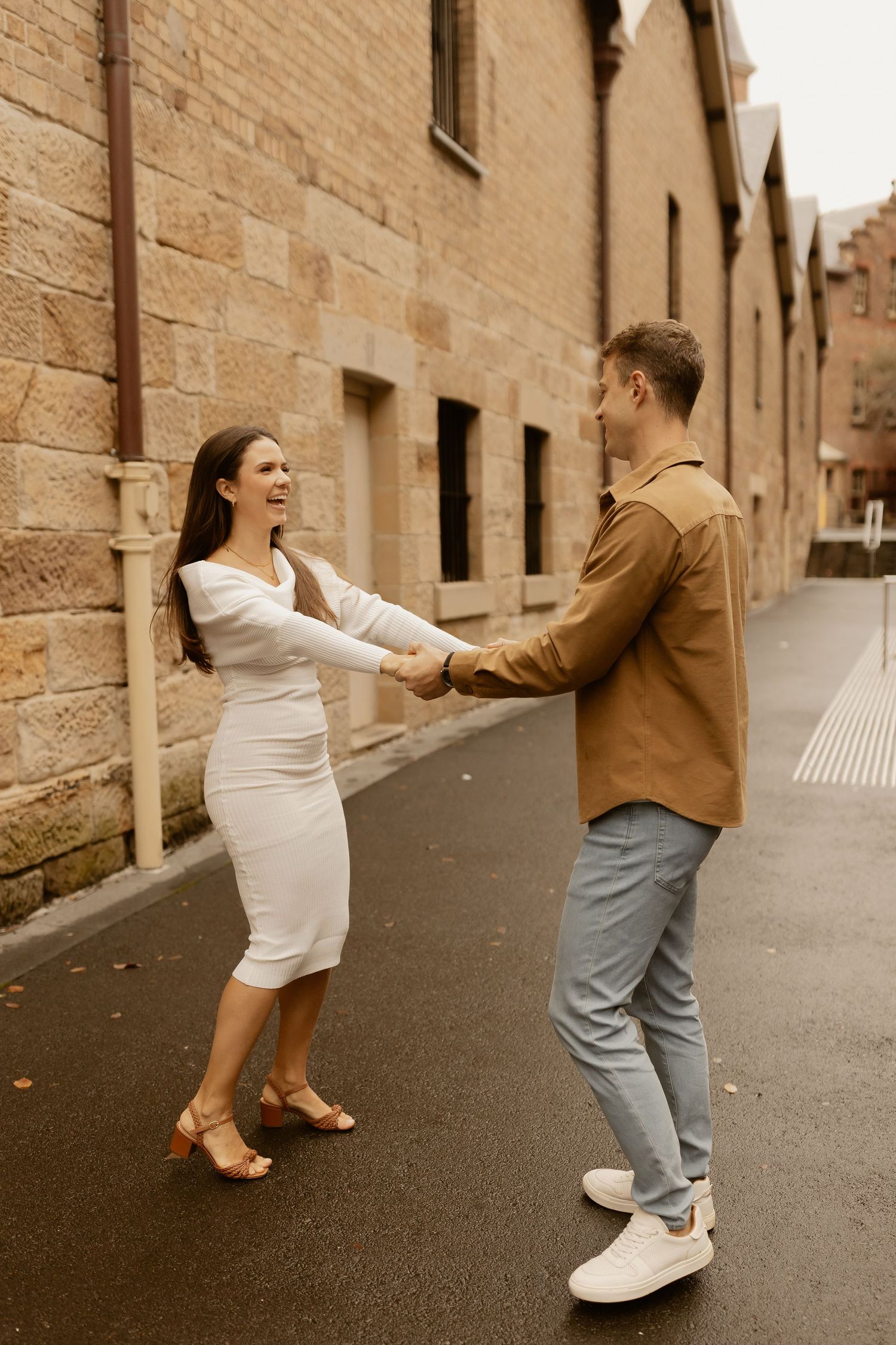 A couple shares a joyful moment dancing together on a street with sandstone buildings in the background.