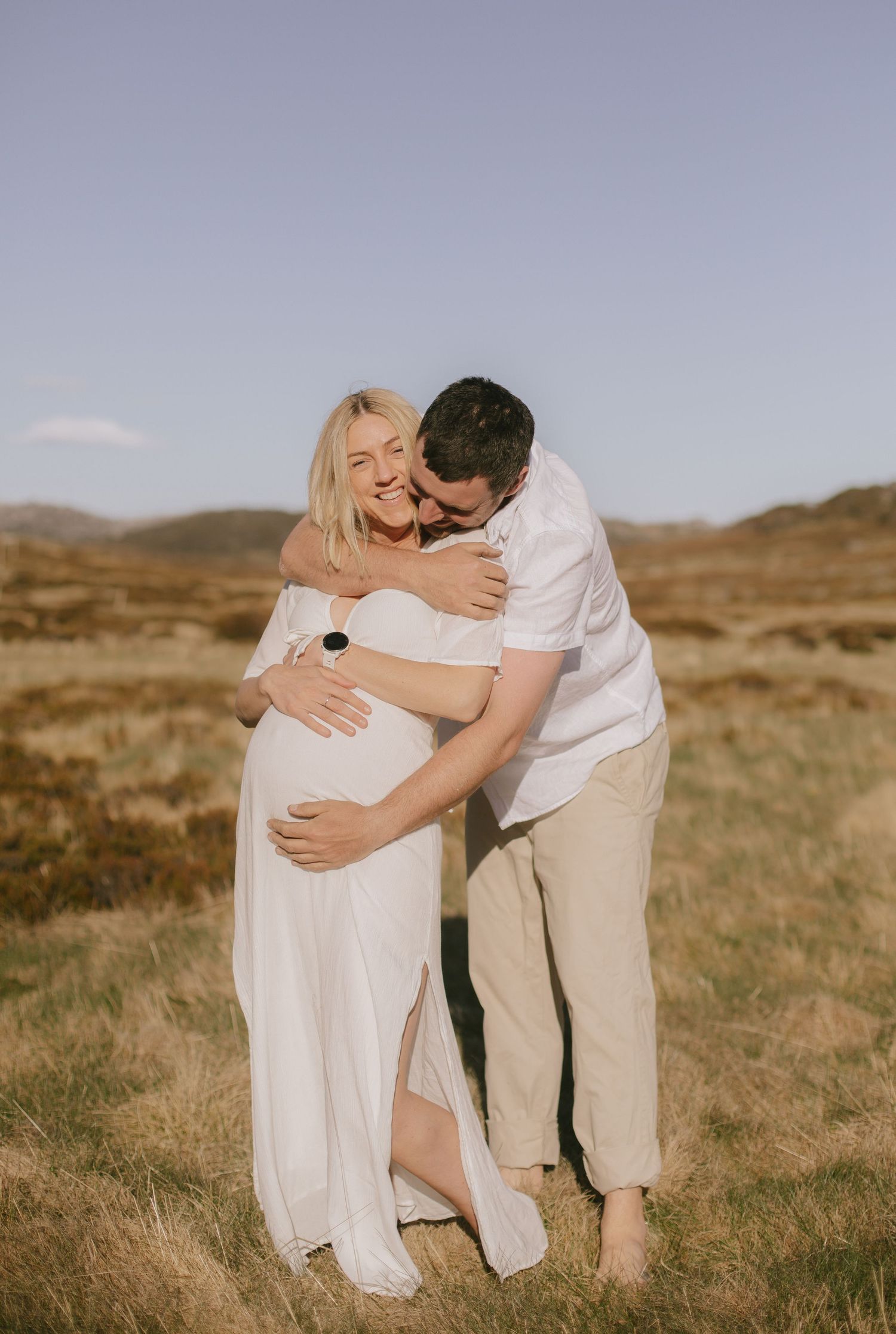 A couple embrace in a flowing white dress and casual attire against a golden rural landscape at sunset.