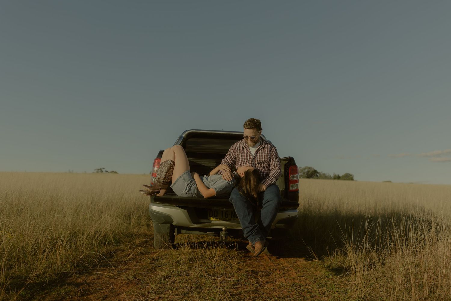 Couple relaxing on tailgate of pickup truck in golden grass field at dusk.