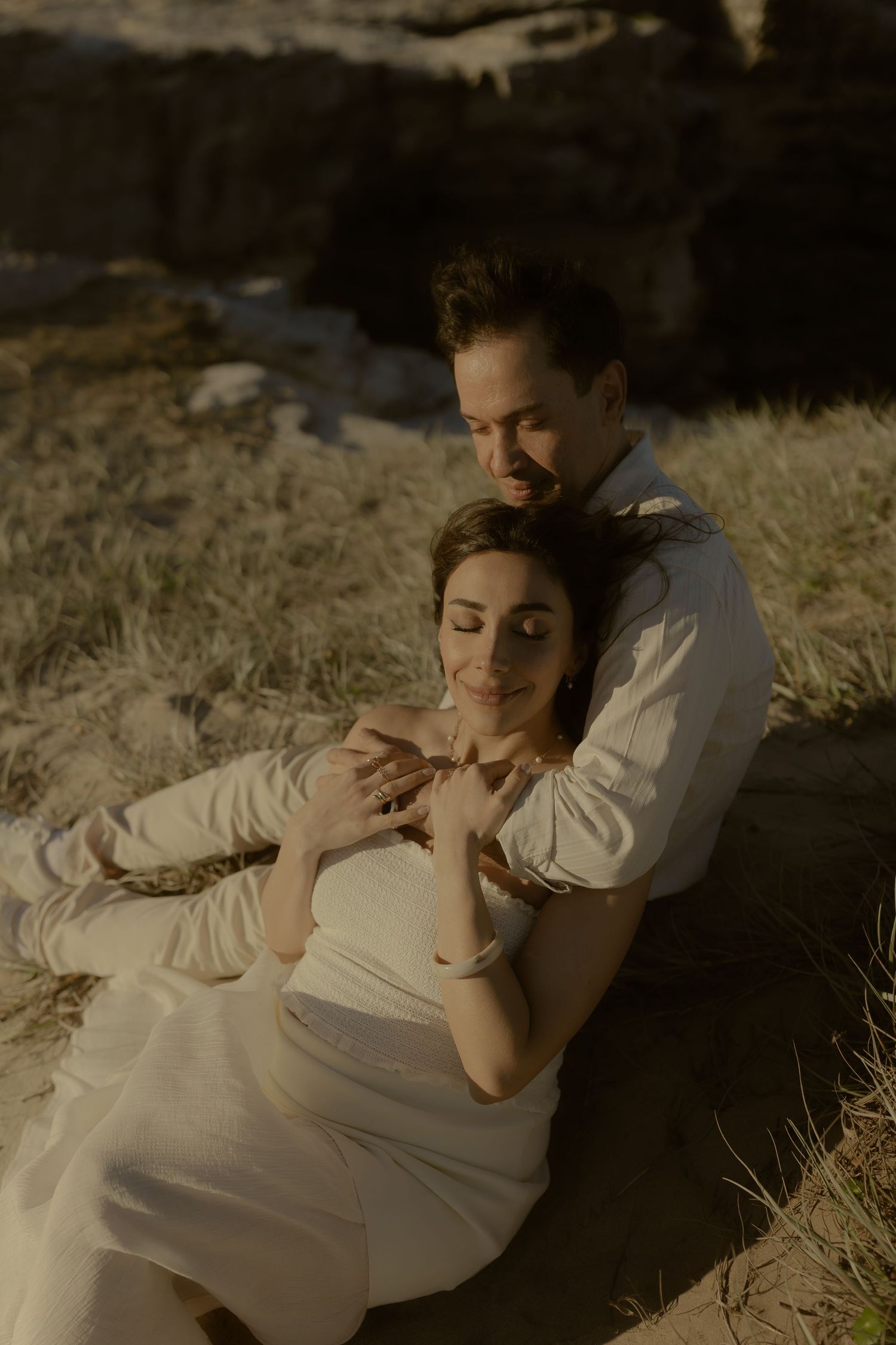 Couple sitting on sandy ground, man embracing pregnant woman from behind in warm golden light.