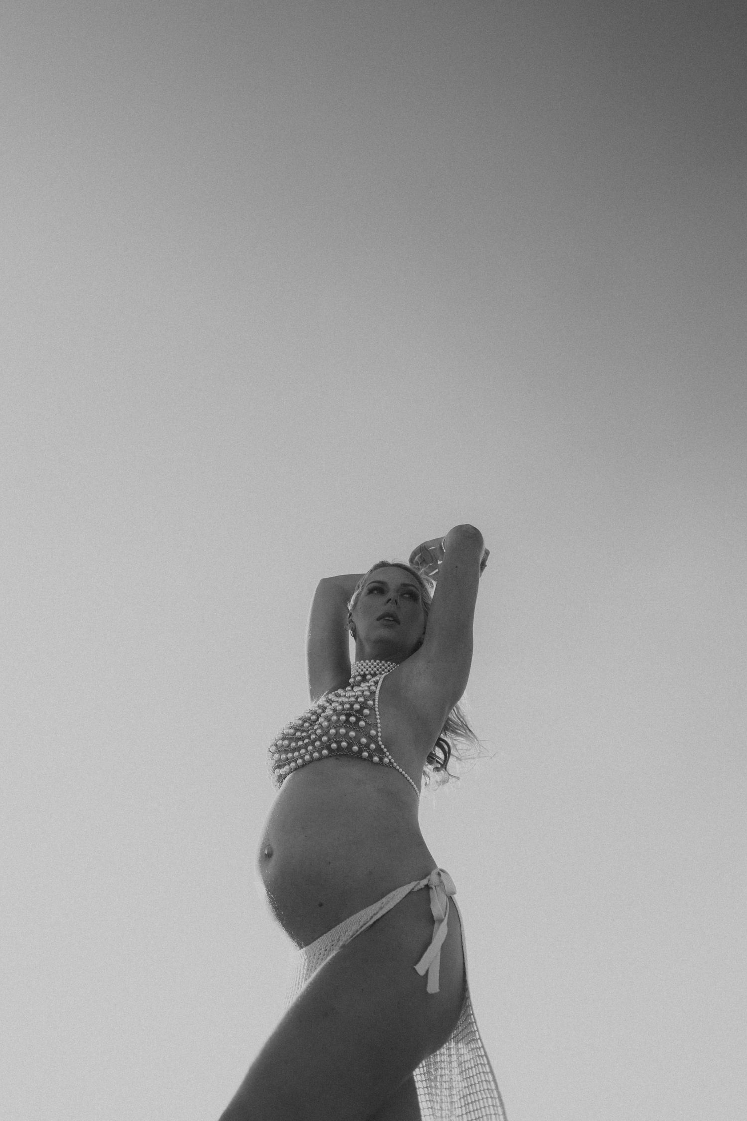 Black and white photo of a pregnant woman in a pearl bikini posing against a clear sky.
