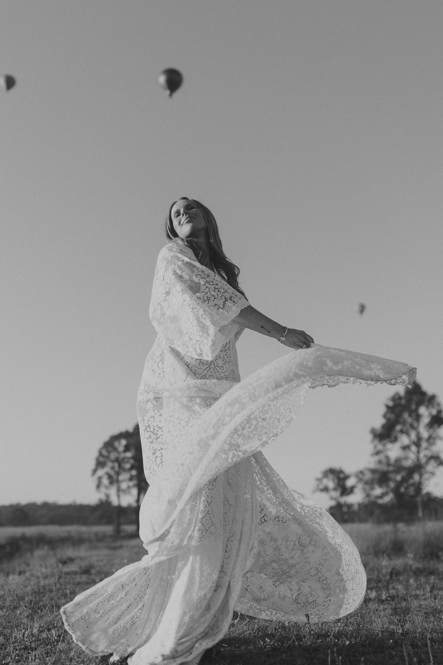 Black and white photo of a bride in lace wedding dress twirling in a field with hot air balloons in the sky.