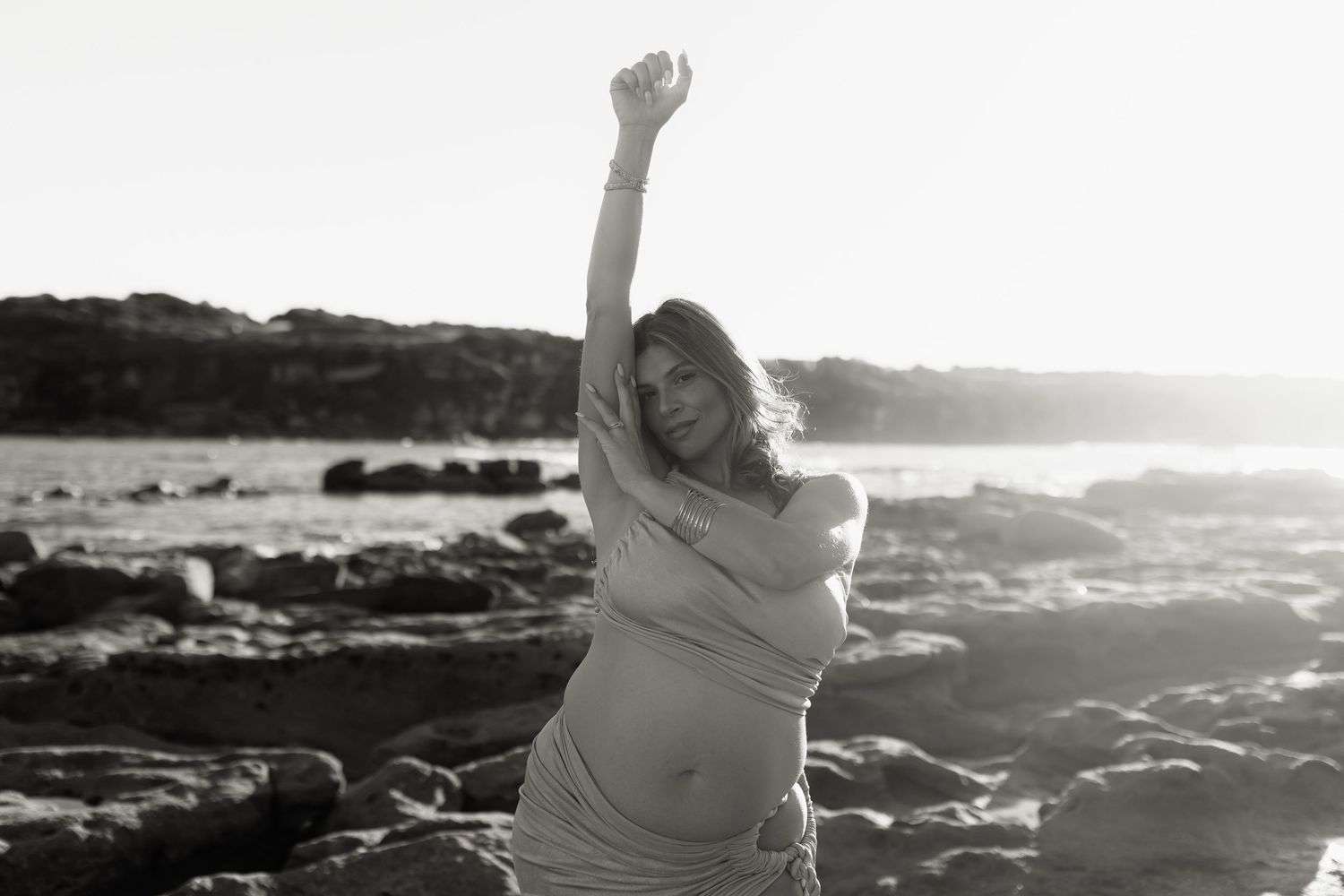 Pregnant woman in flowing wrap poses on rocky beach, arm raised, in artistic black and white maternity photo.