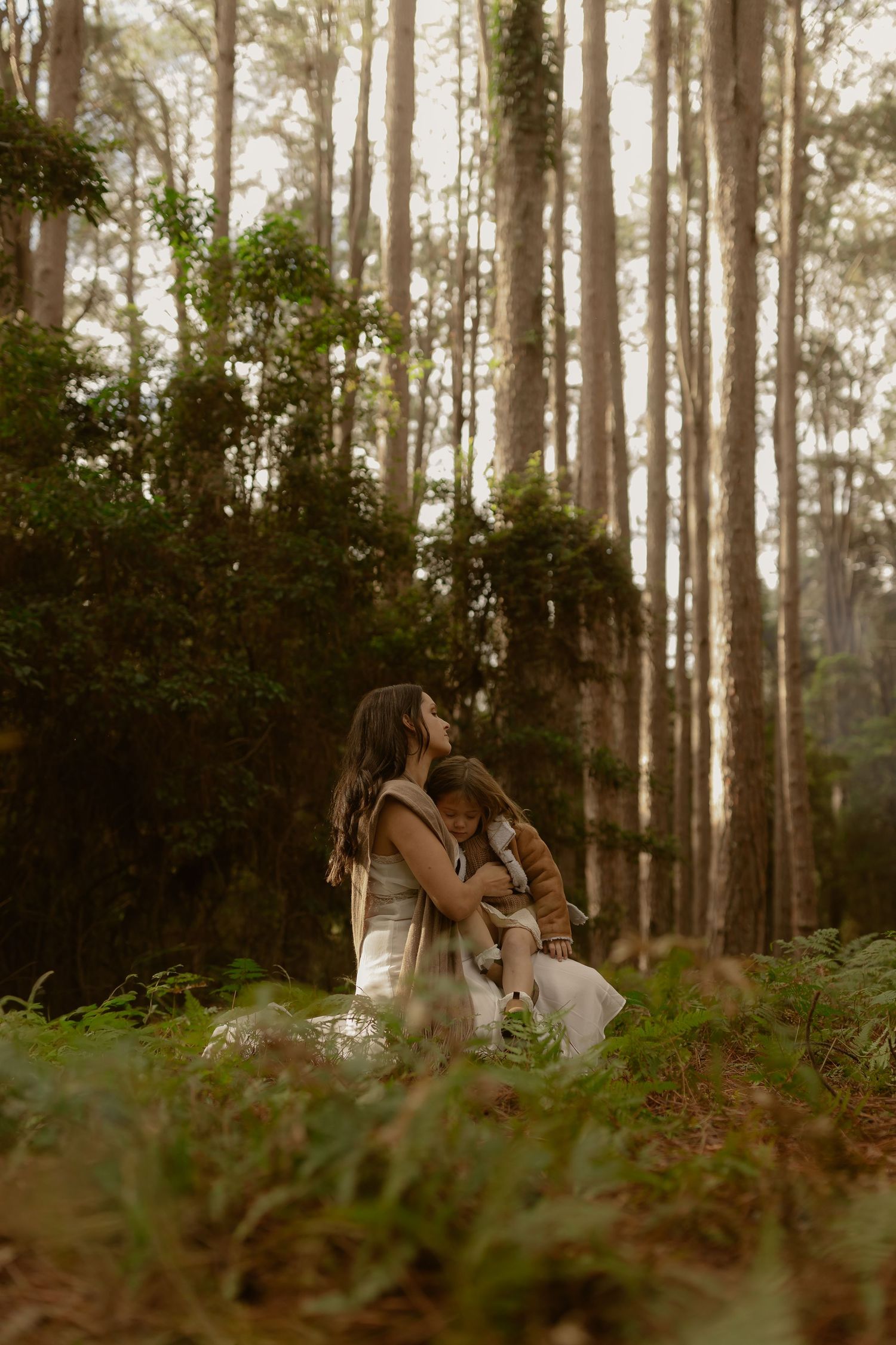 Mother crouches to kiss young child among ferns in a sunlit forest.