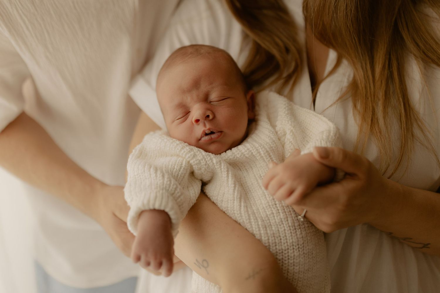 A sleeping newborn baby wearing white lies peacefully on soft white bedding while being gently held.