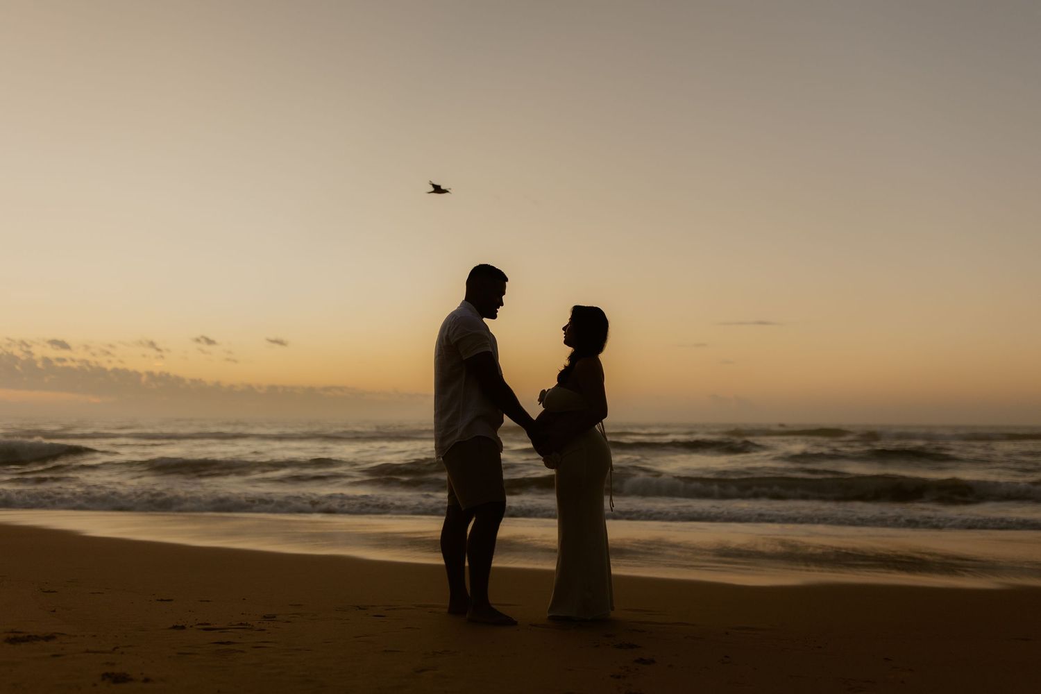 Two silhouettes stand together on a beach at sunset with waves crashing in the background.