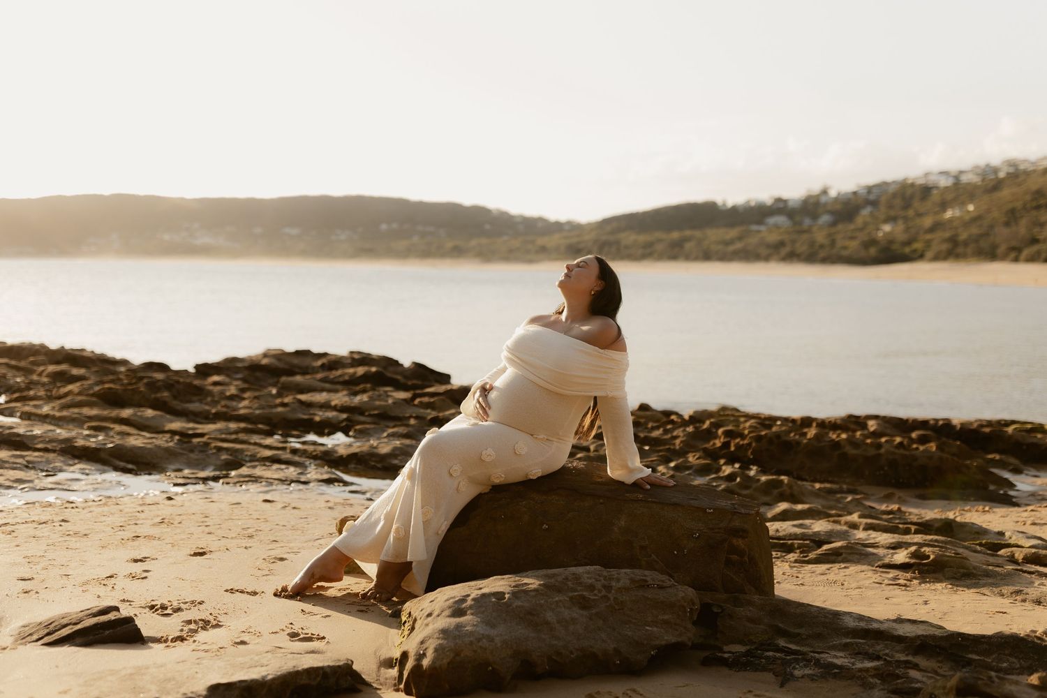 A couple in white wedding attire sits together on coastal rocks overlooking a serene ocean view at sunset.