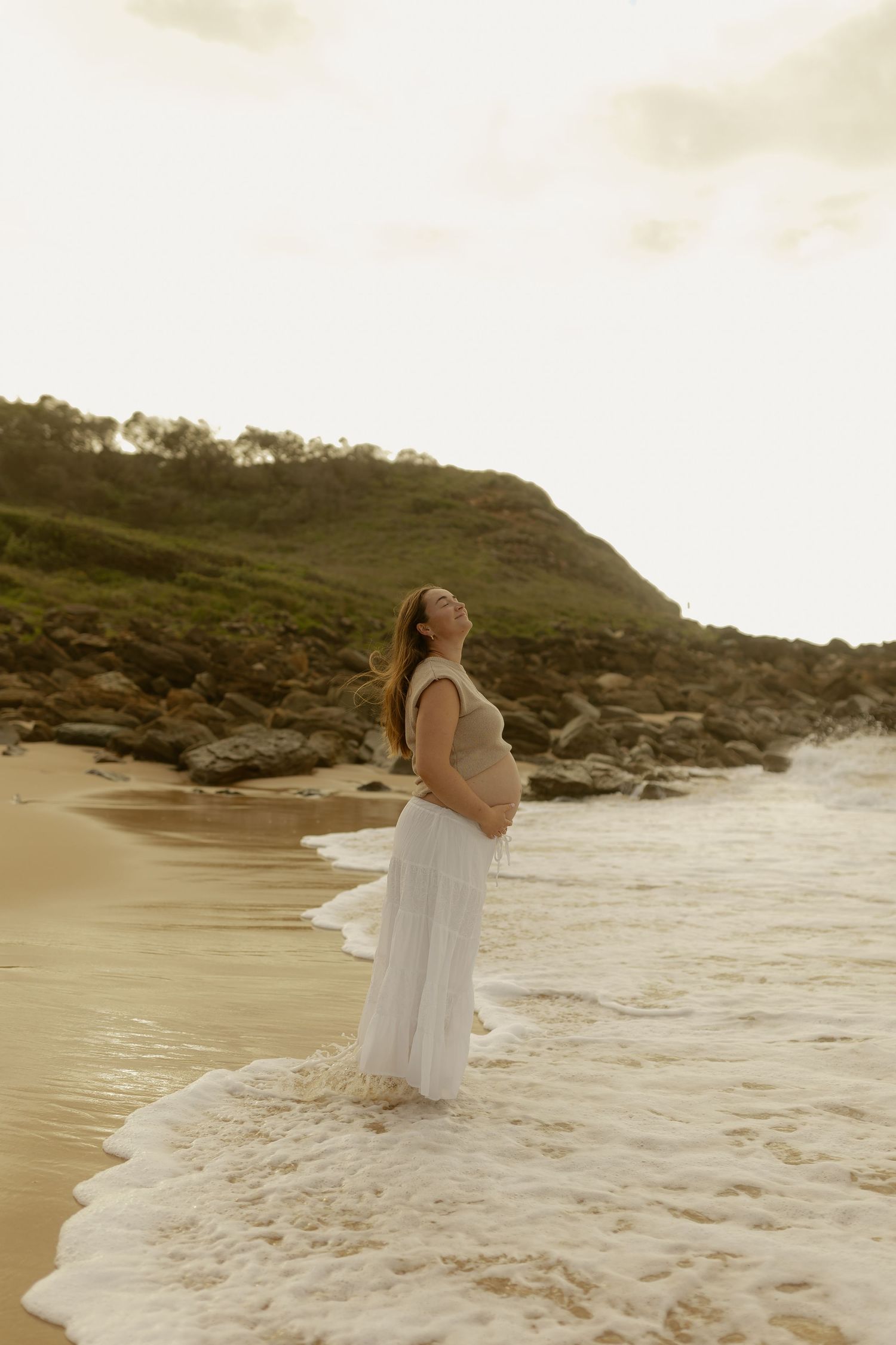 A person in a white dress stands in the ocean waves at sunset on a rocky beach.