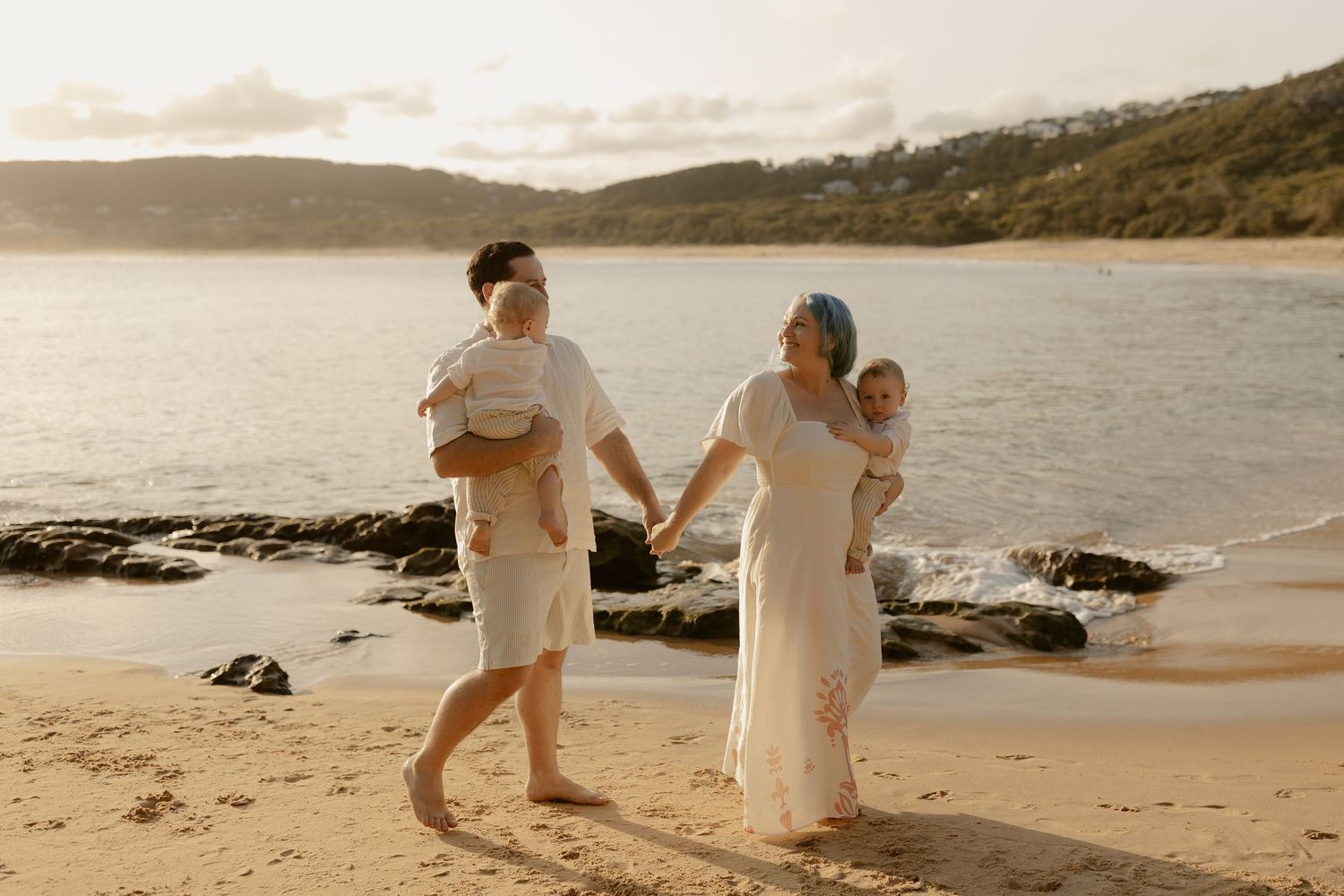 Family walking along a scenic beach at sunset with rocky coastline and mountains in the background.