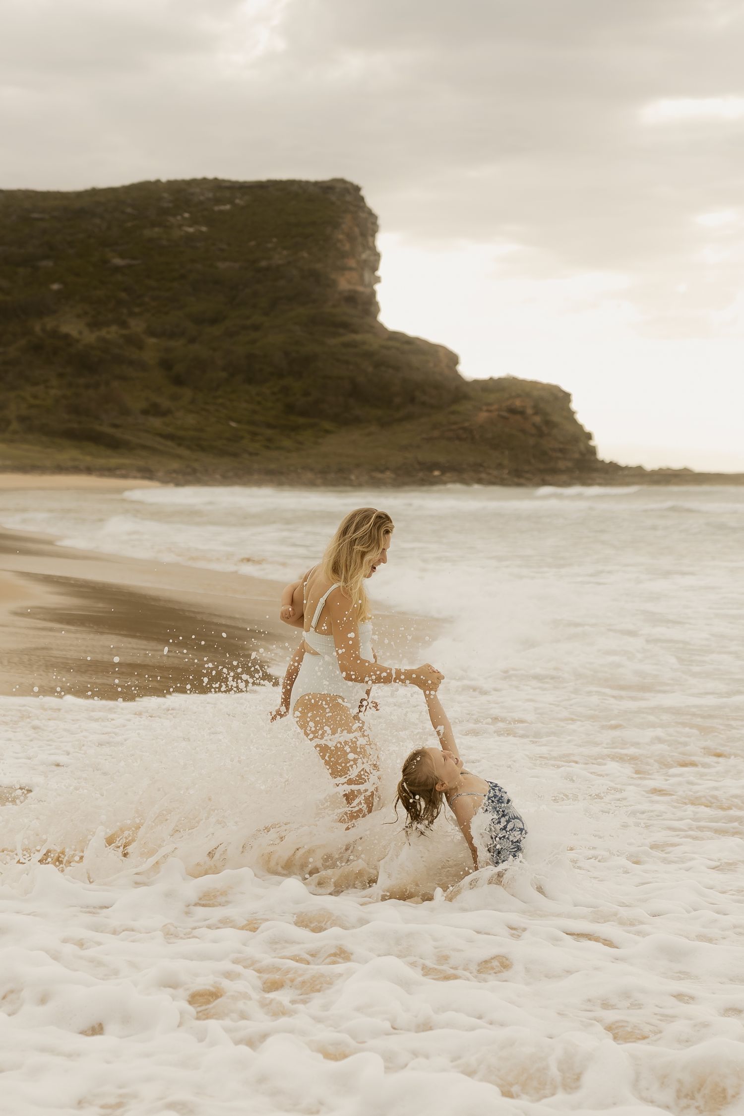 Two people playing in crashing ocean waves near dramatic coastal cliffs at sunset.