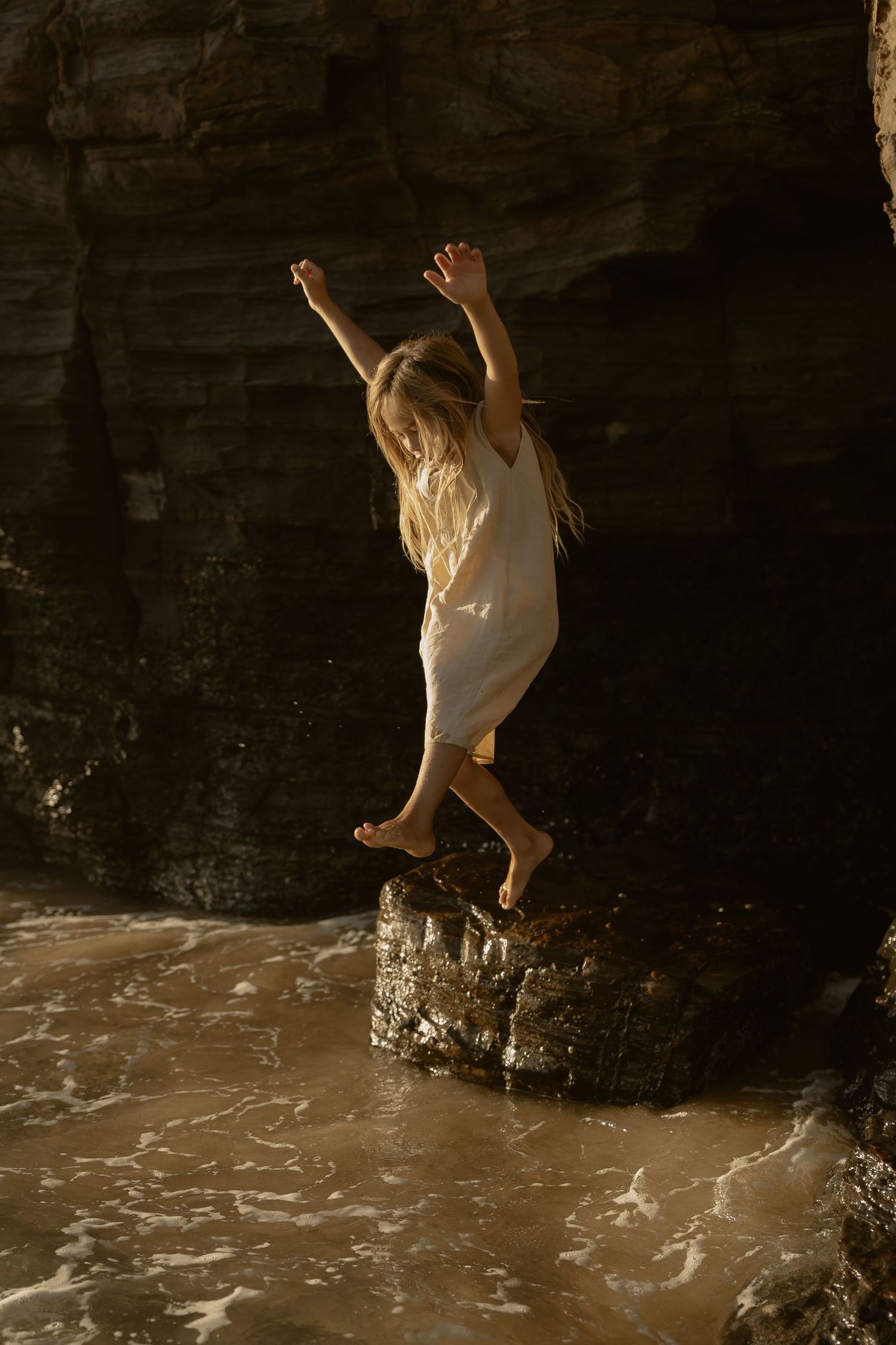 Child in white dress jumping joyfully off a rock formation at the beach during sunset.