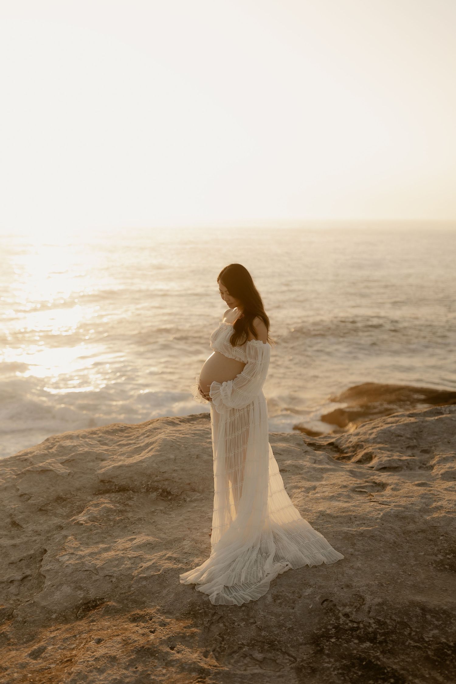 A figure in a flowing white dress stands gracefully on rocky coastal cliffs at sunset by the ocean.