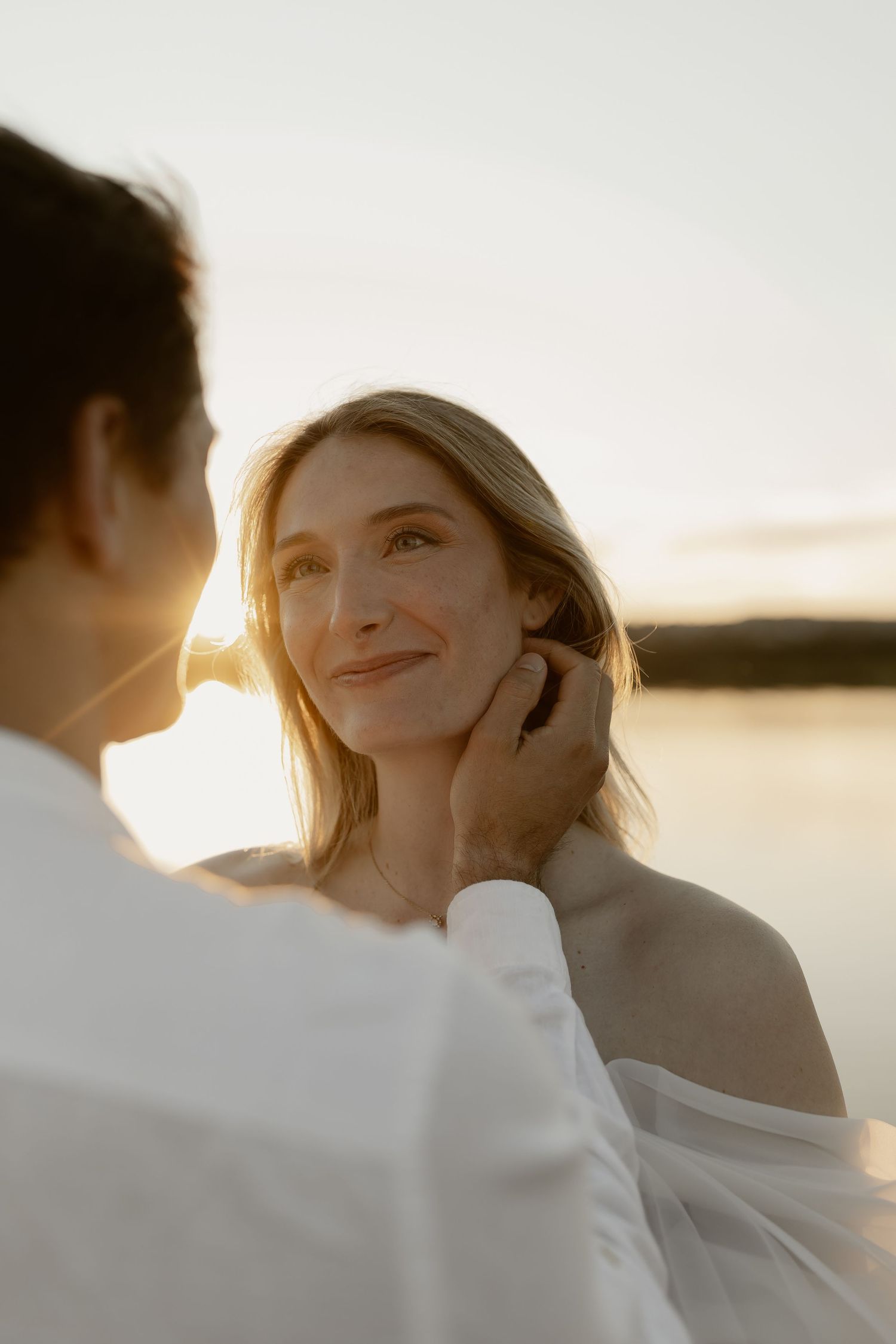 A couple shares an intimate moment at sunset by the water, with soft golden light illuminating their silhouettes.
