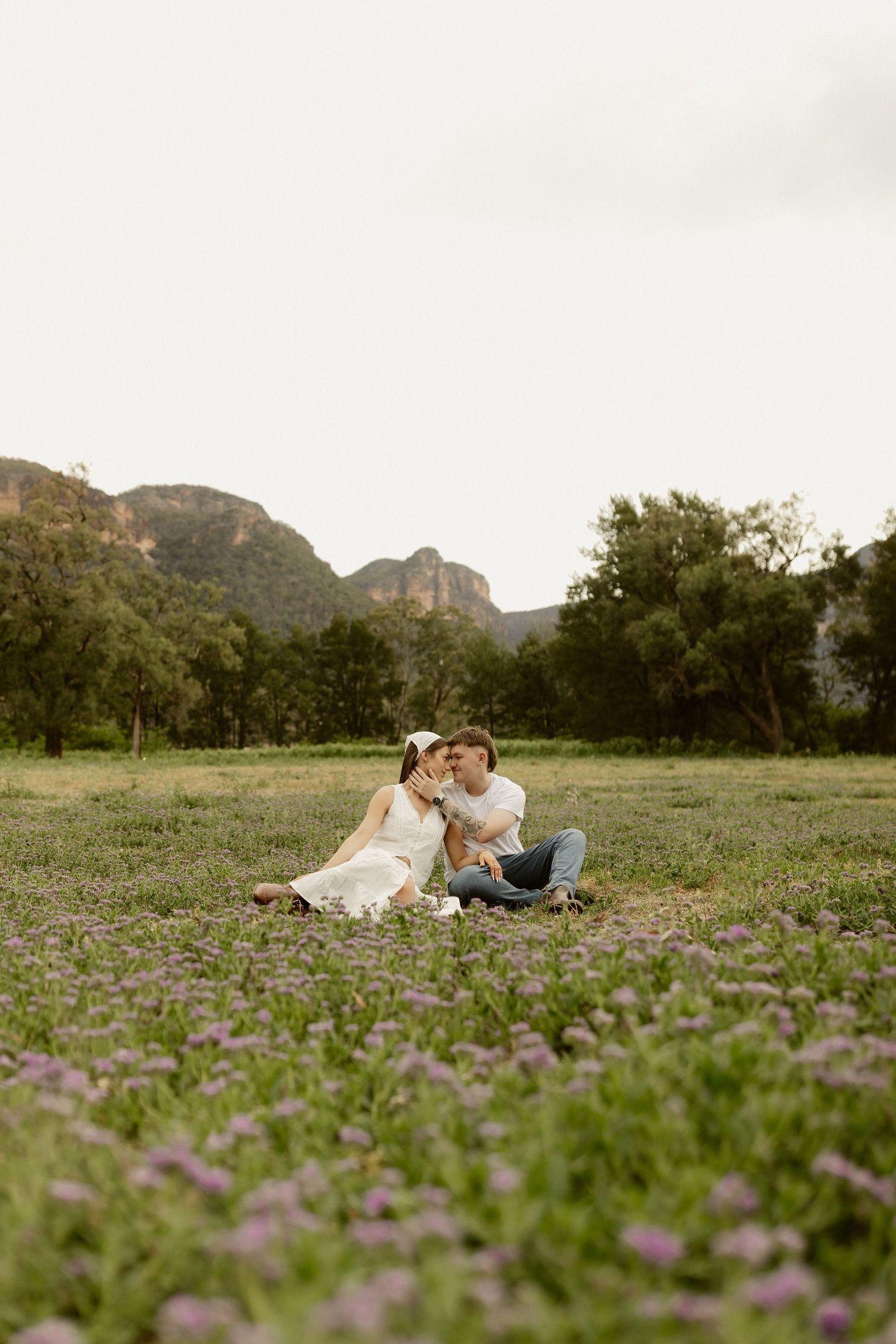 A couple sits together in a field of purple wildflowers with mountains in the background.