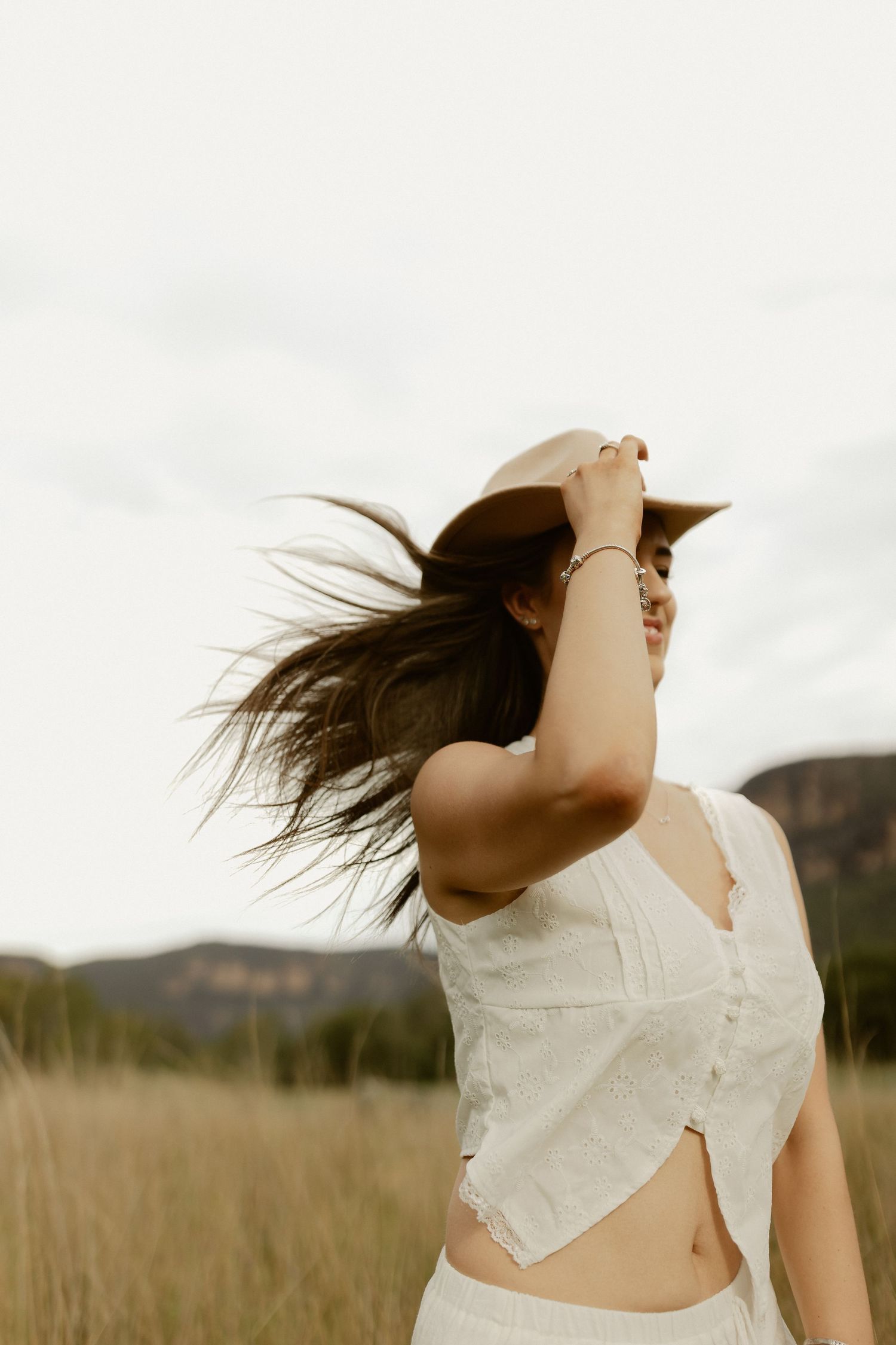 Woman in white top with flowing hair against cloudy sky in countryside setting.
