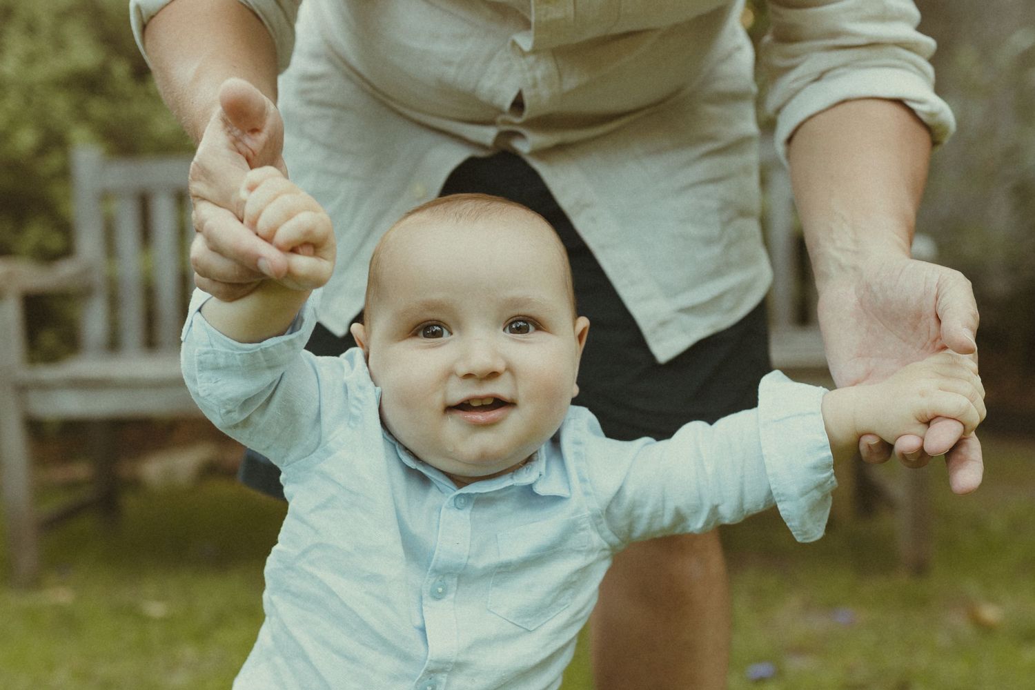Baby in light blue outfit reaching up playfully while being held outdoors.