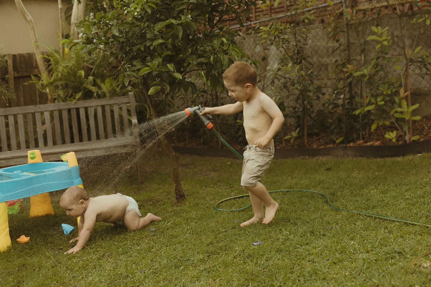 Children playing with a water sprinkler in a backyard with toys on a summer day.