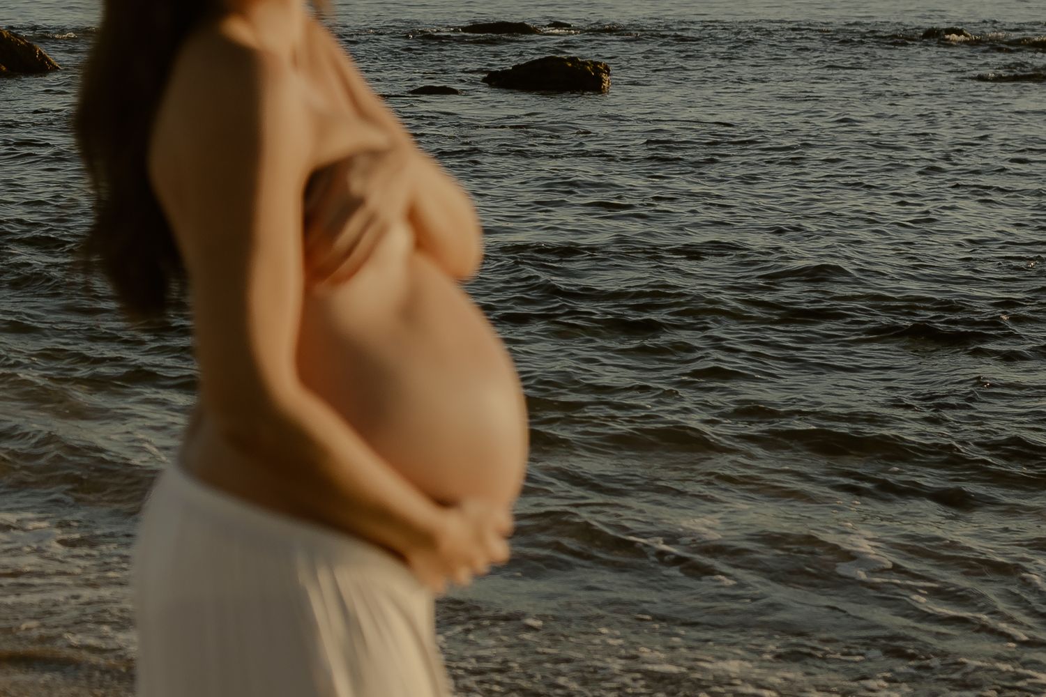 A pregnant woman in a white dress stands near dark ocean waves at sunset, cradling her belly.