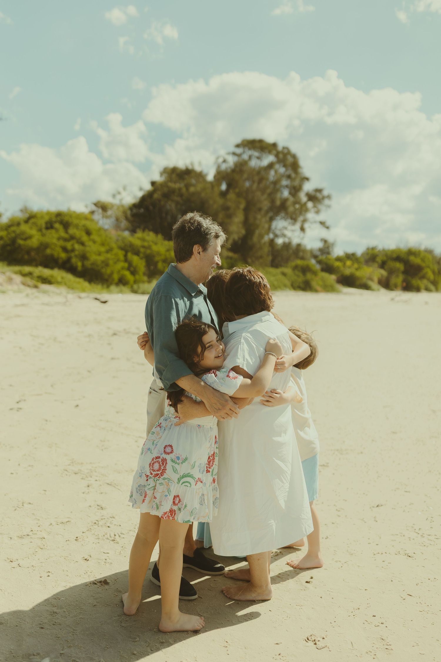 Intimate family embrace captured on a sunlit sandy beach with coastal vegetation.