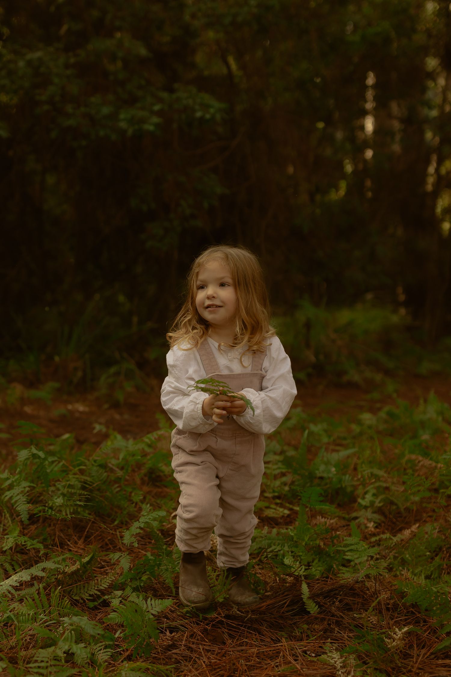 A young blonde girl in white top and linen overalls stands in a forest, holding a green leaf, smiling softly.