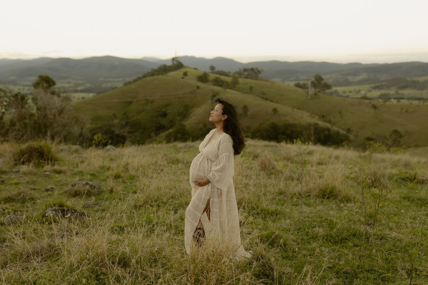 Pregnant woman in white dress standing in green rolling hills landscape during golden hour.