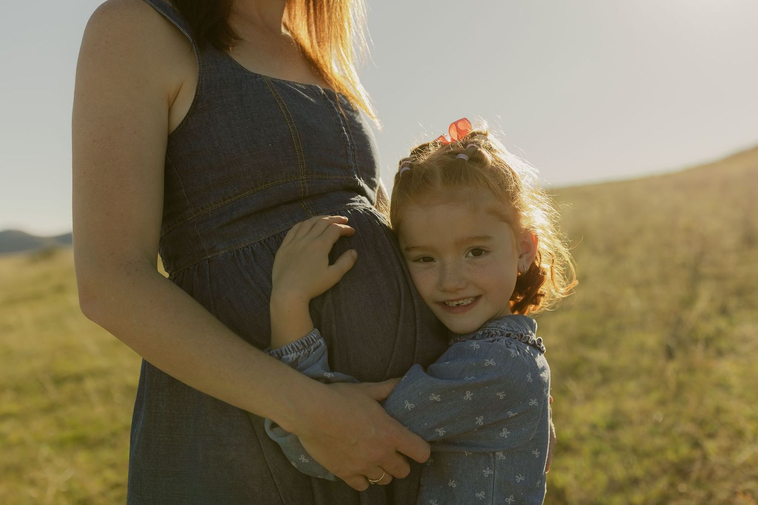 Young girl hugging pregnant mother's belly outdoors in sunlit green field, sweet maternity moment.