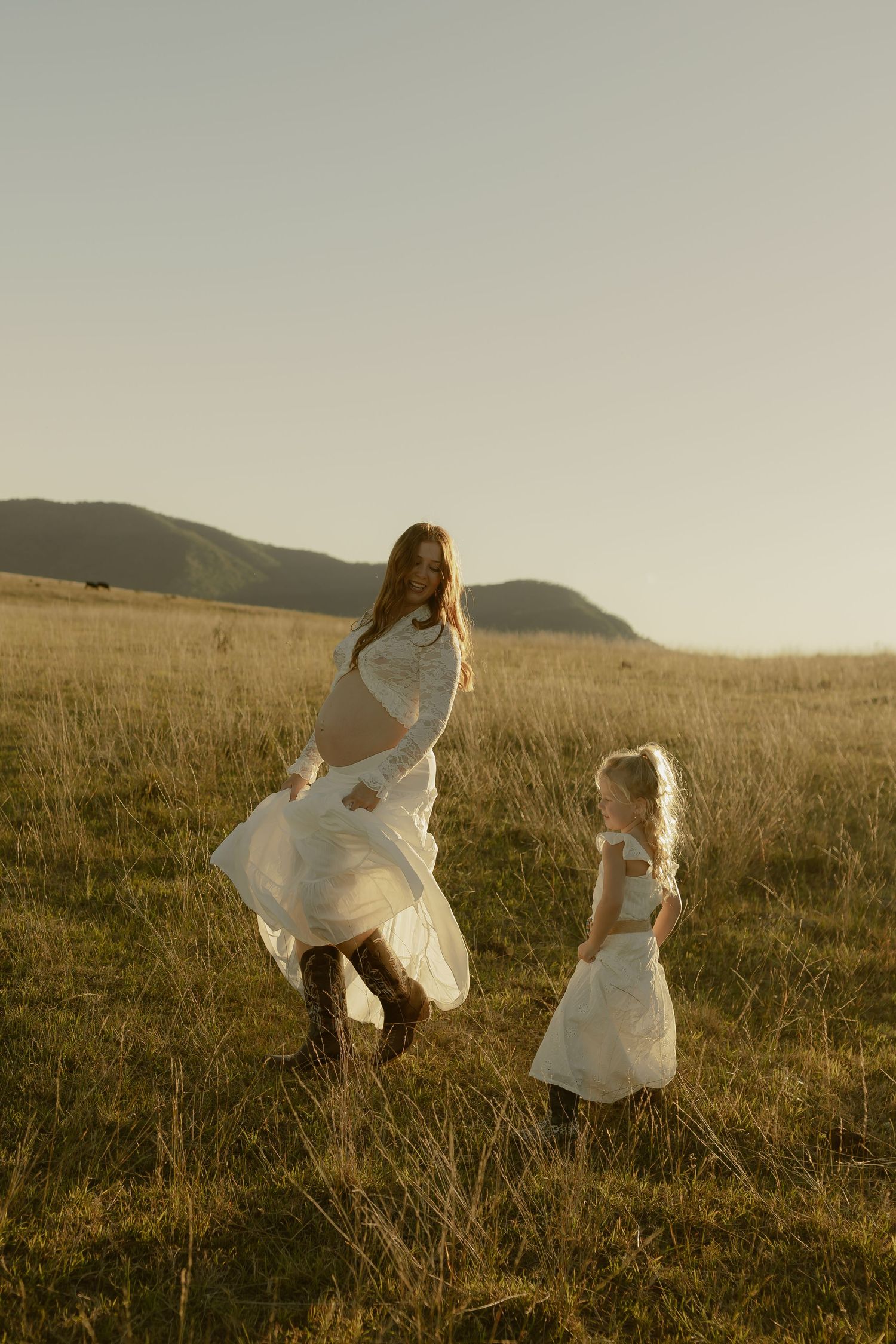 Mother and daughter in white dresses dancing in golden field with rolling hills at sunset.