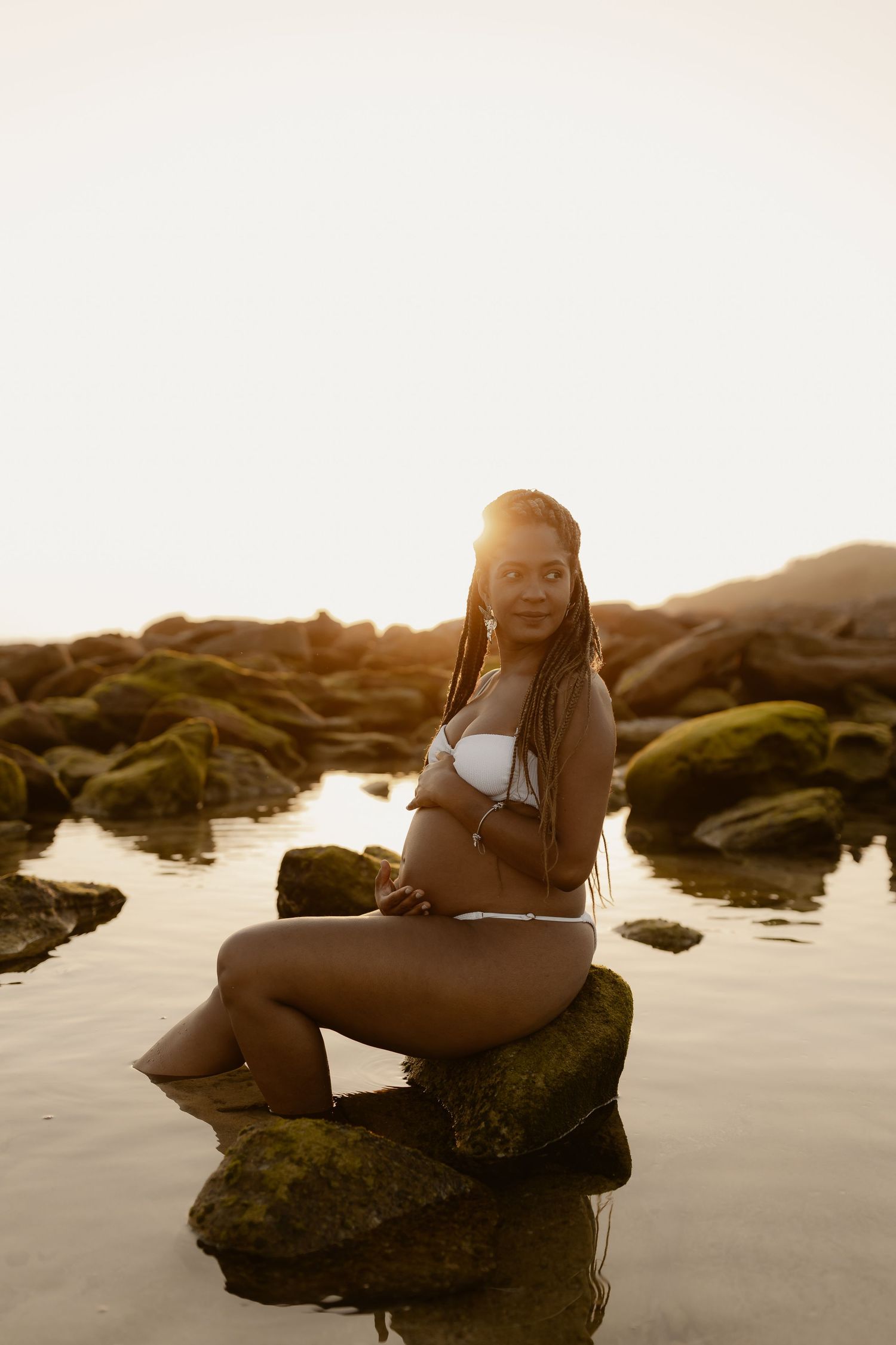 A person poses on rocky beach formations during golden hour, with gentle waves and mossy rocks in the background.