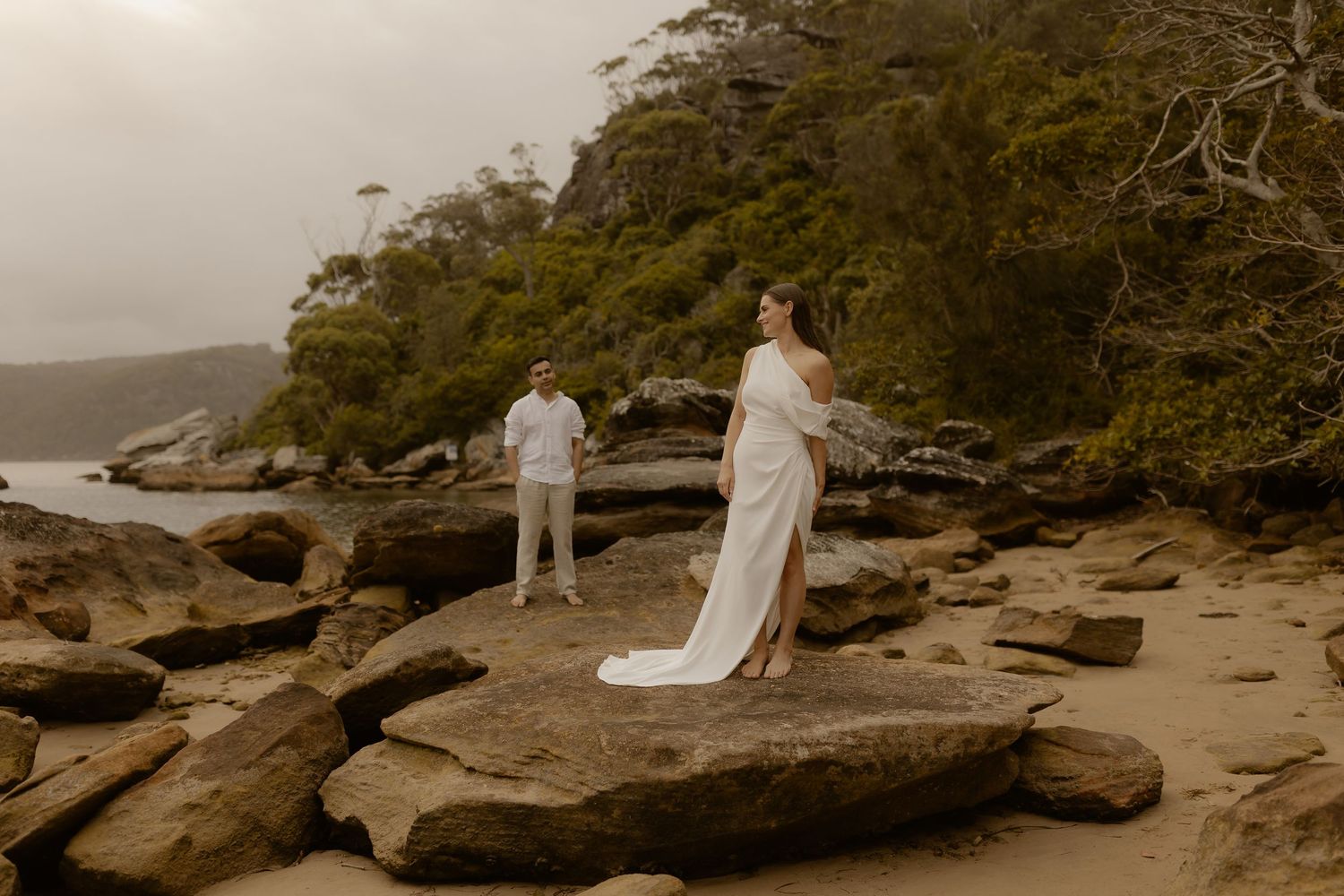A couple in white attire poses on rocky coastal terrain with trees and water in the background.