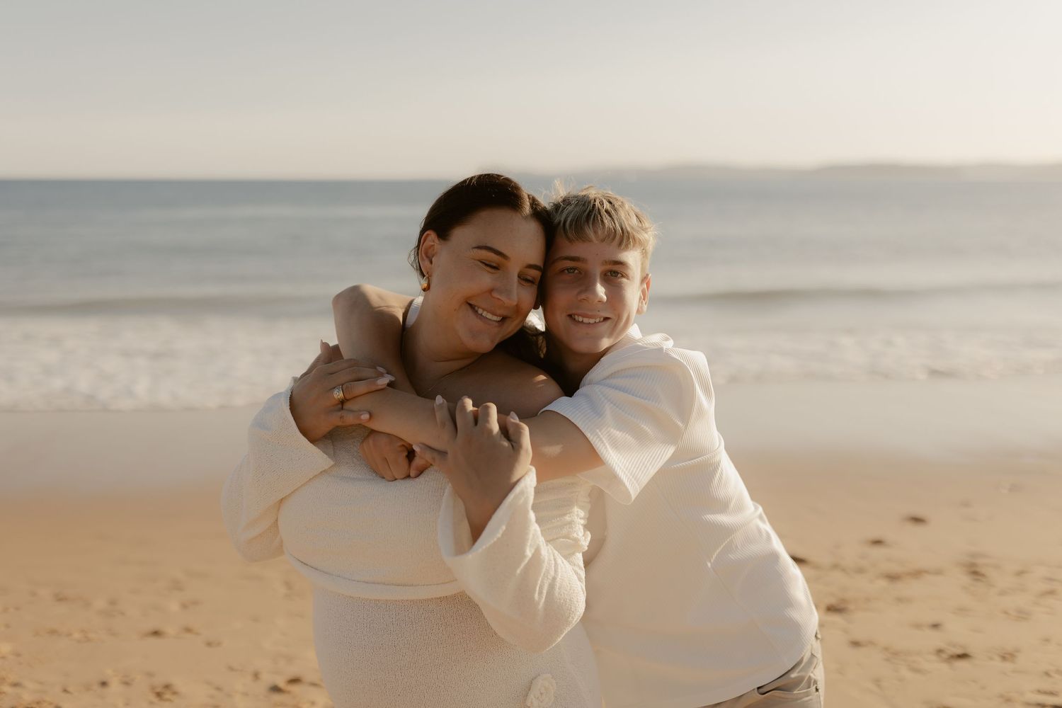 Two people in white outfits embrace while sharing a joyful moment on a sunny beach with ocean in background.