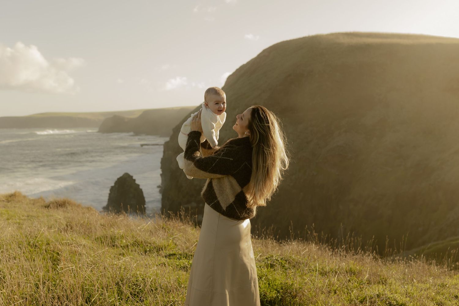 A mother lifts her baby joyfully on a scenic coastal cliff at golden hour, with ocean waves and green hills in the background.