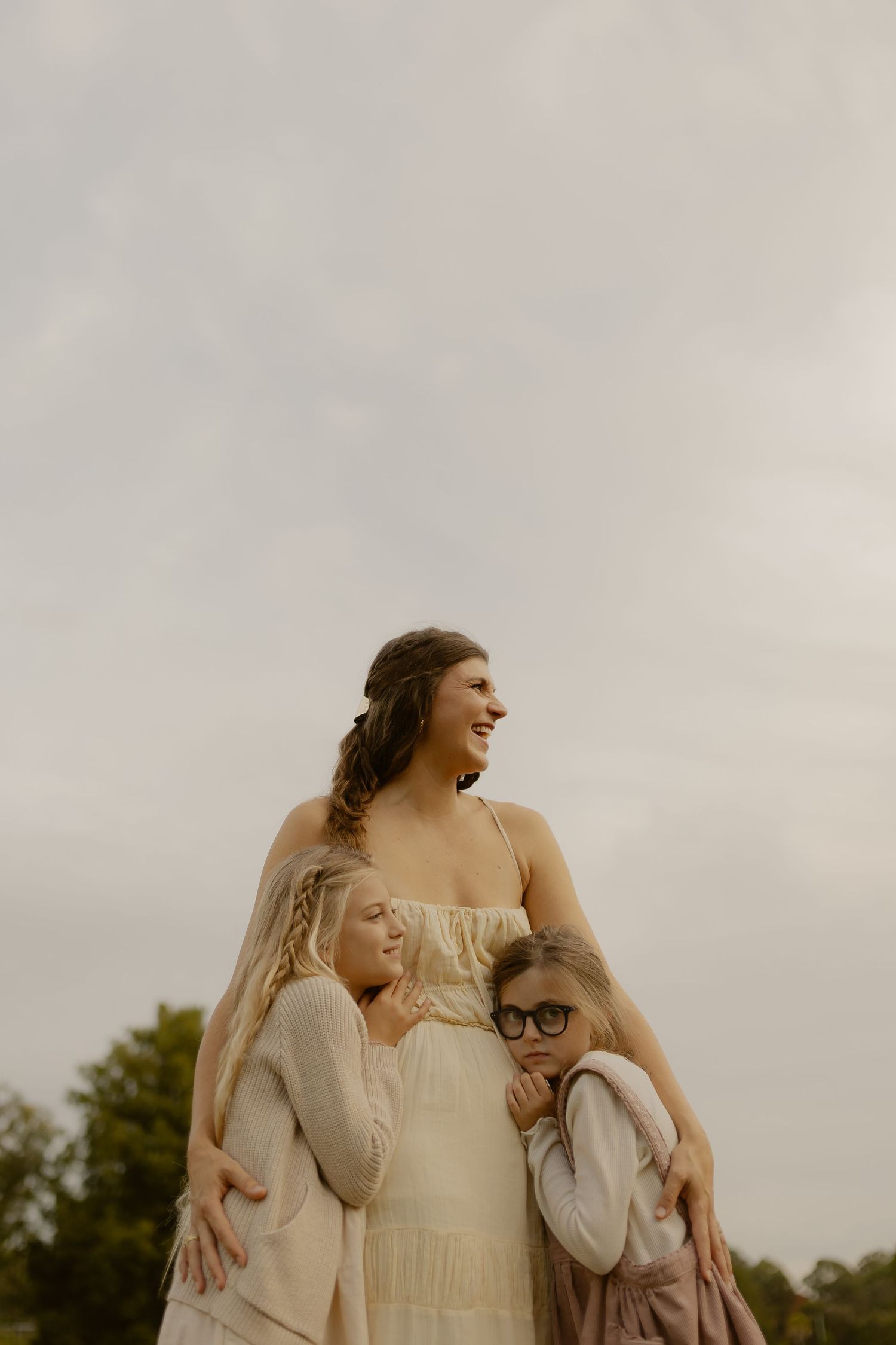 A smiling mother in a beige dress embraces her two daughters under a soft cloudy sky outdoors.