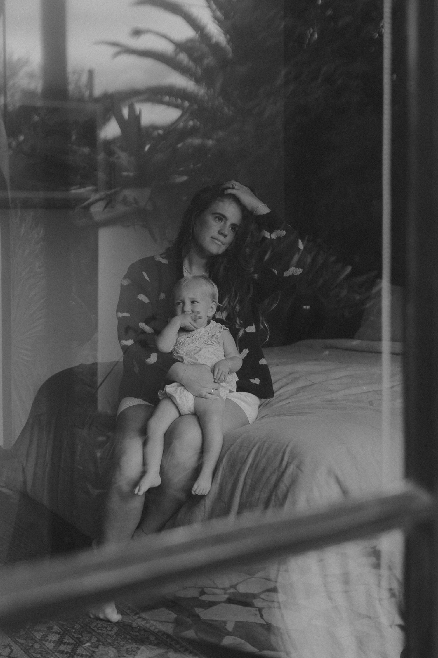 Black and white photo of mother holding baby on bed, shot through window with palm trees behind.