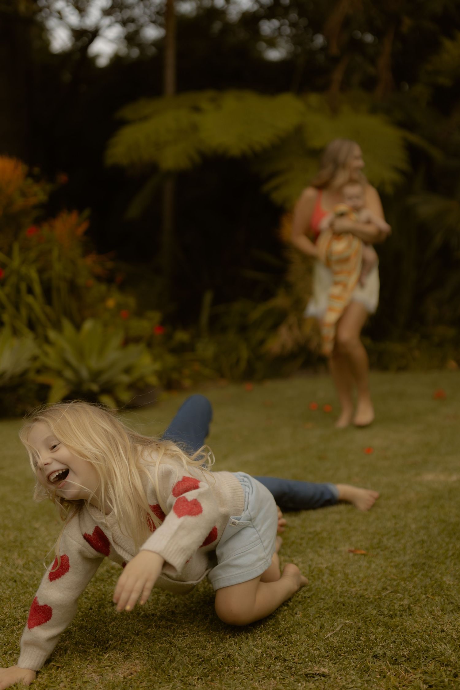 A laughing blonde girl crawls on green grass as a woman holds a baby in a garden background.