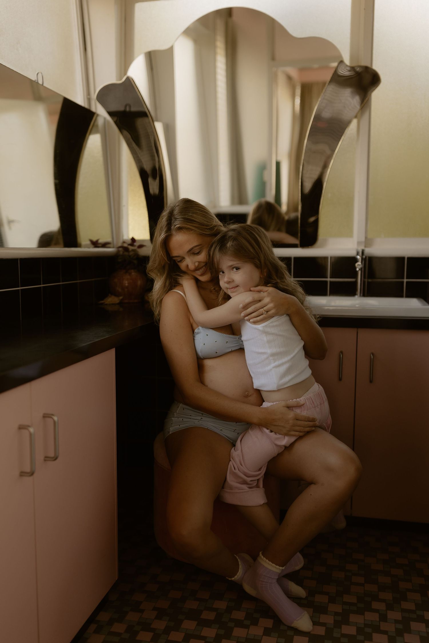 Pregnant woman hugging young daughter in vintage pink bathroom with decorative mirrors.