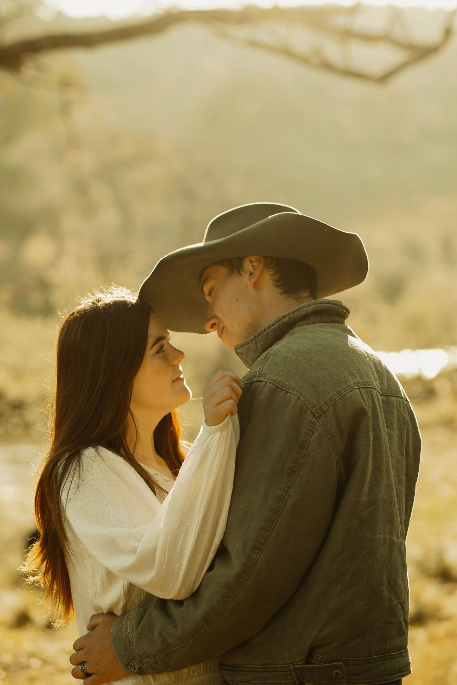 A romantic couple embraces in golden sunlight, with the person in a cowboy hat and denim jacket against a dreamy backdrop.