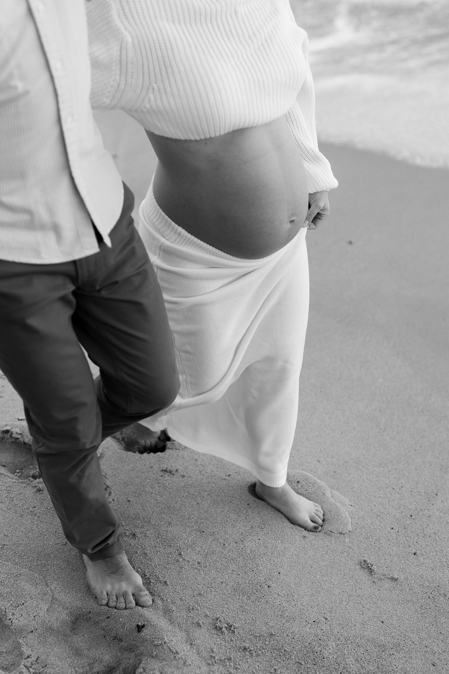 Close-up black and white photo of bare feet on sand and flowing white maternity dress.