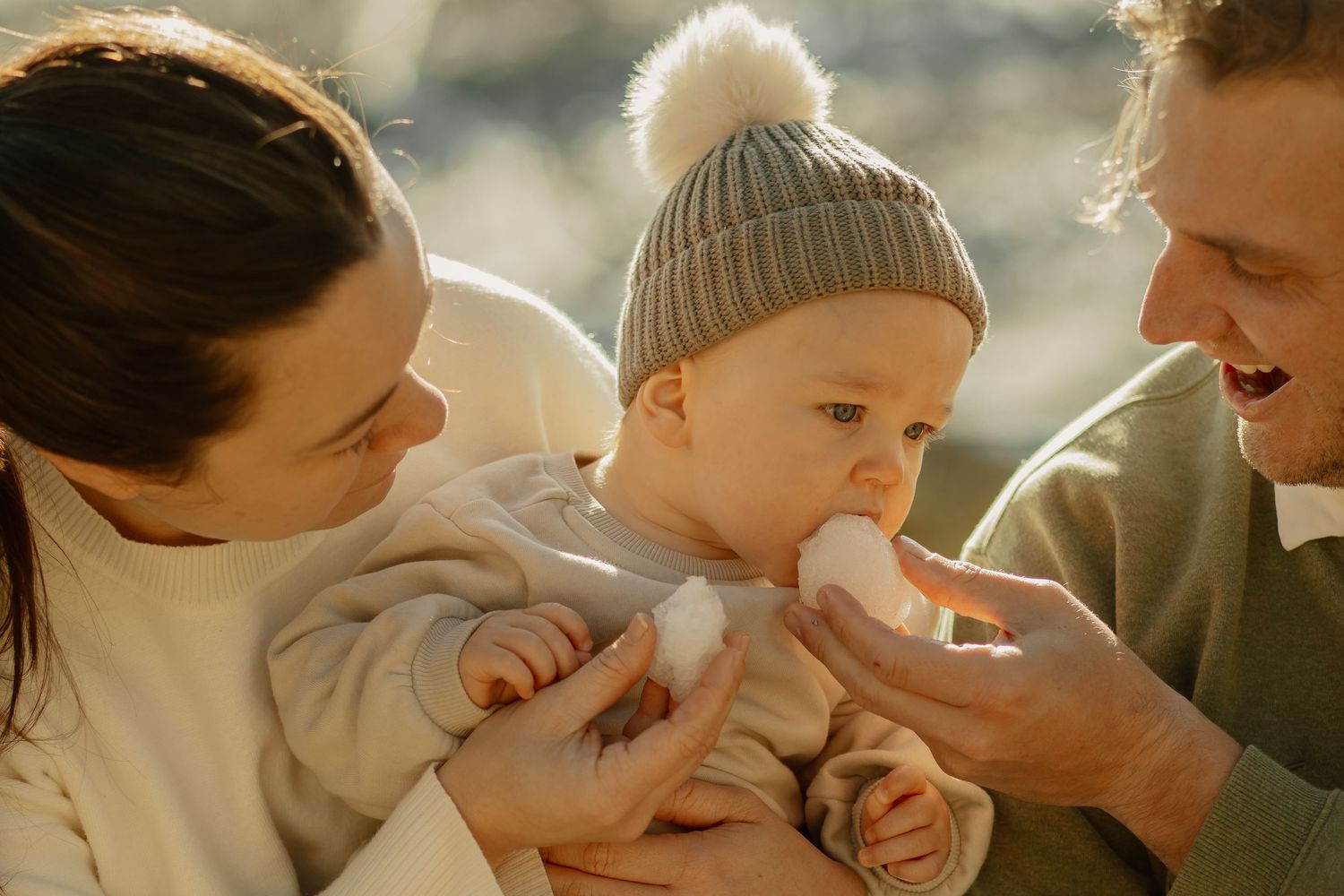A baby in a gray winter hat is being bottle-fed while seated between two adults in warm sunlight.