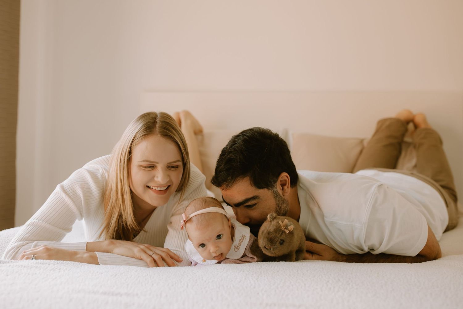 A family lies together on a white bed with their baby, creating a warm and intimate moment.