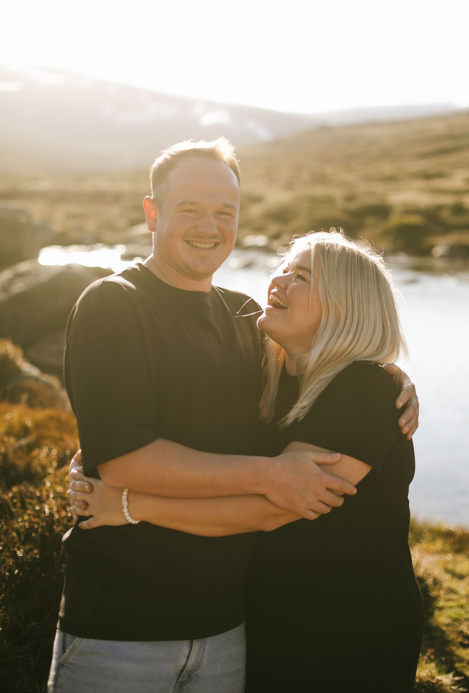 A loving couple embraces and laughs together in golden sunlight with a scenic mountainous landscape in the background.