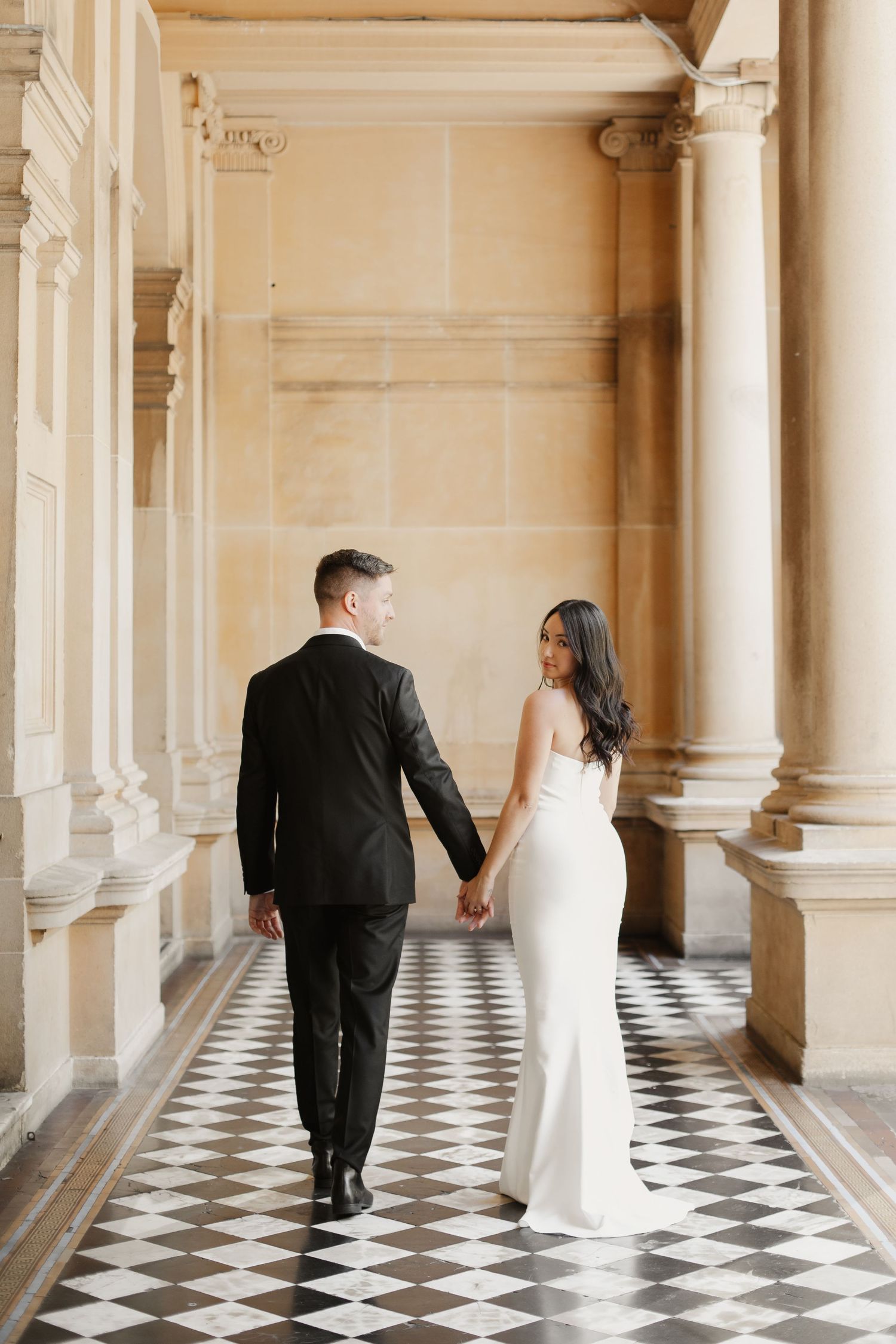 A couple in wedding attire holds hands while walking down an elegant hallway with checkered floors and ornate columns.