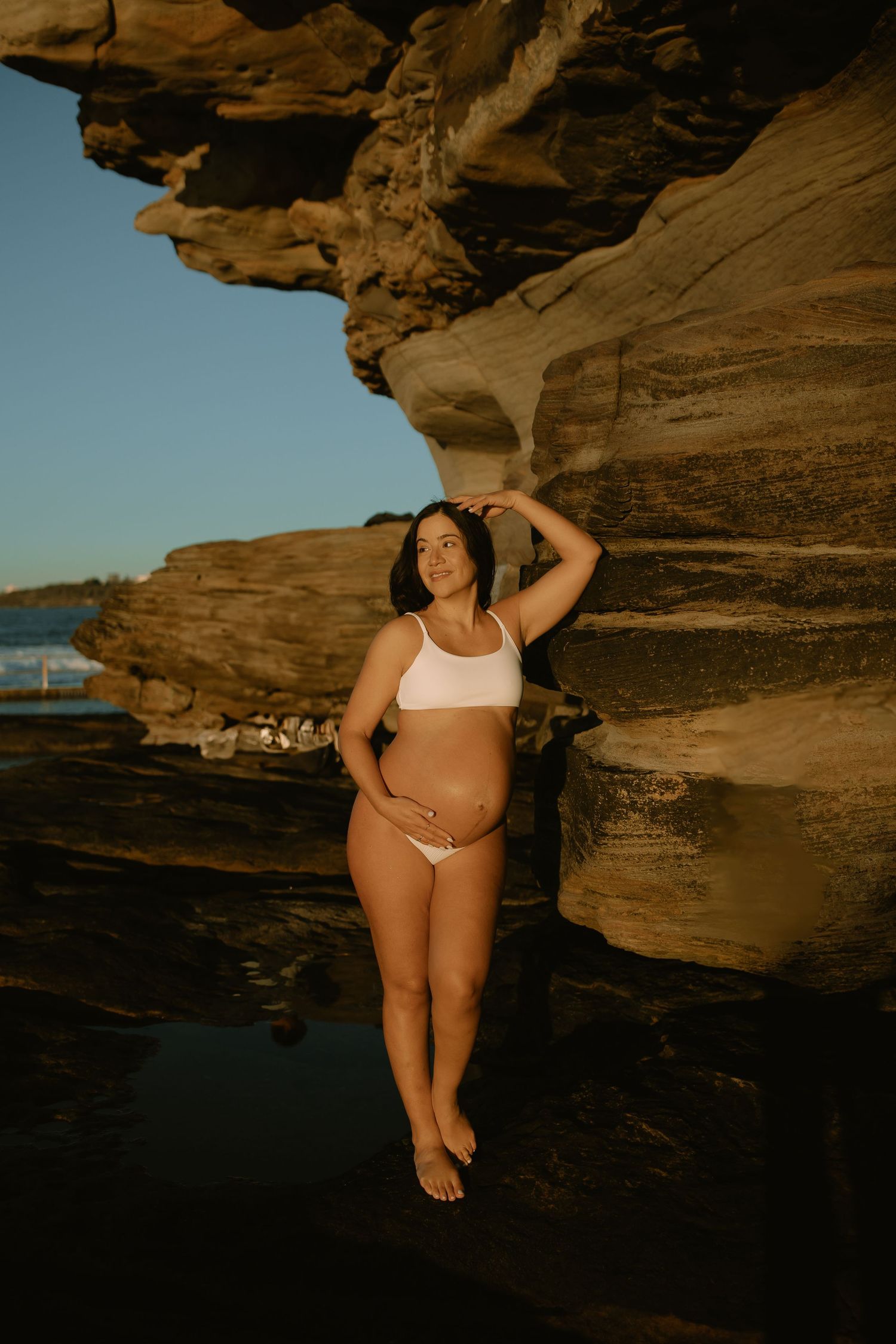 A person poses on rocky coastal cliffs during golden hour in swimwear.