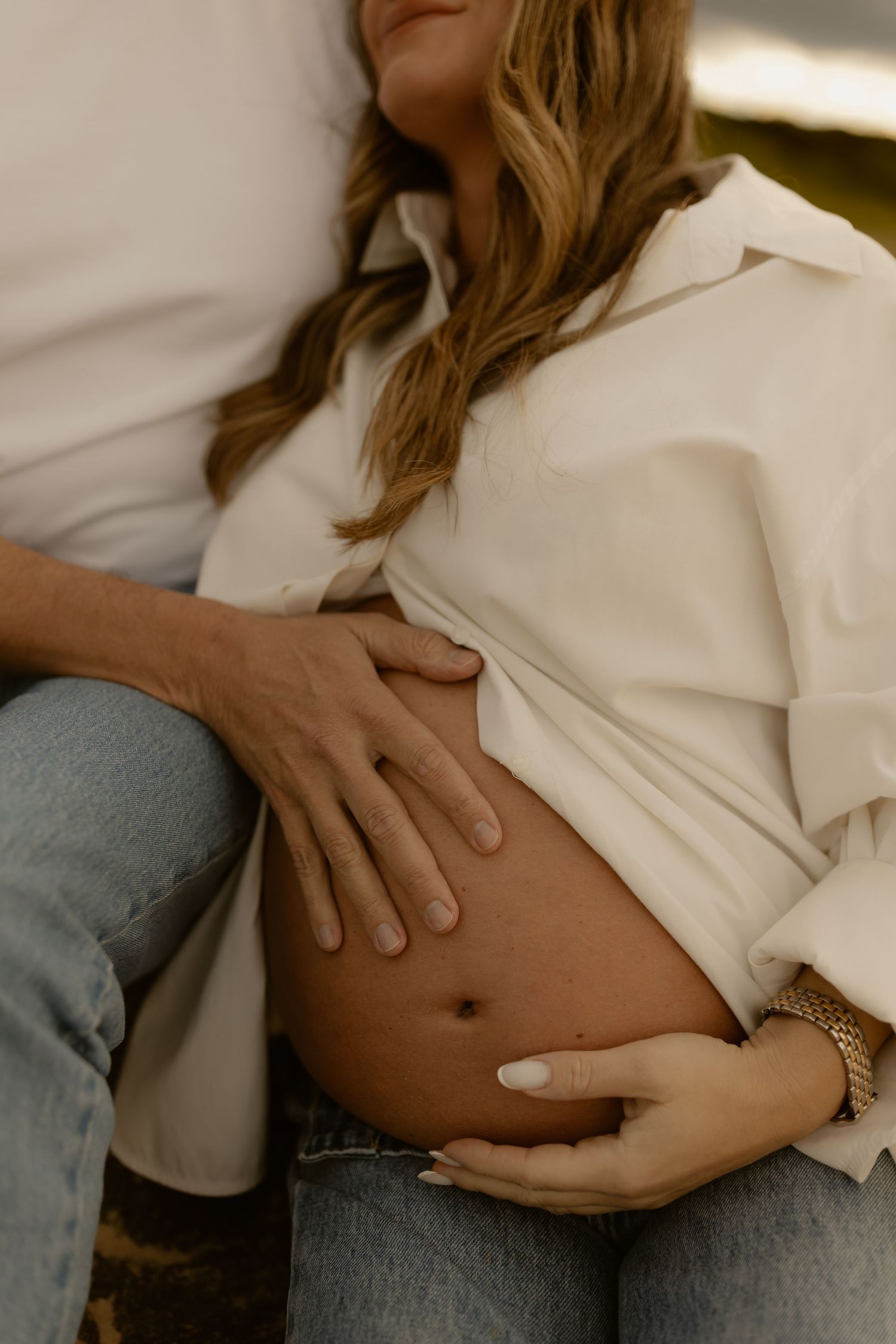 Pregnant woman in white shirt and jeans cradling baby bump with partner's hand resting on belly.
