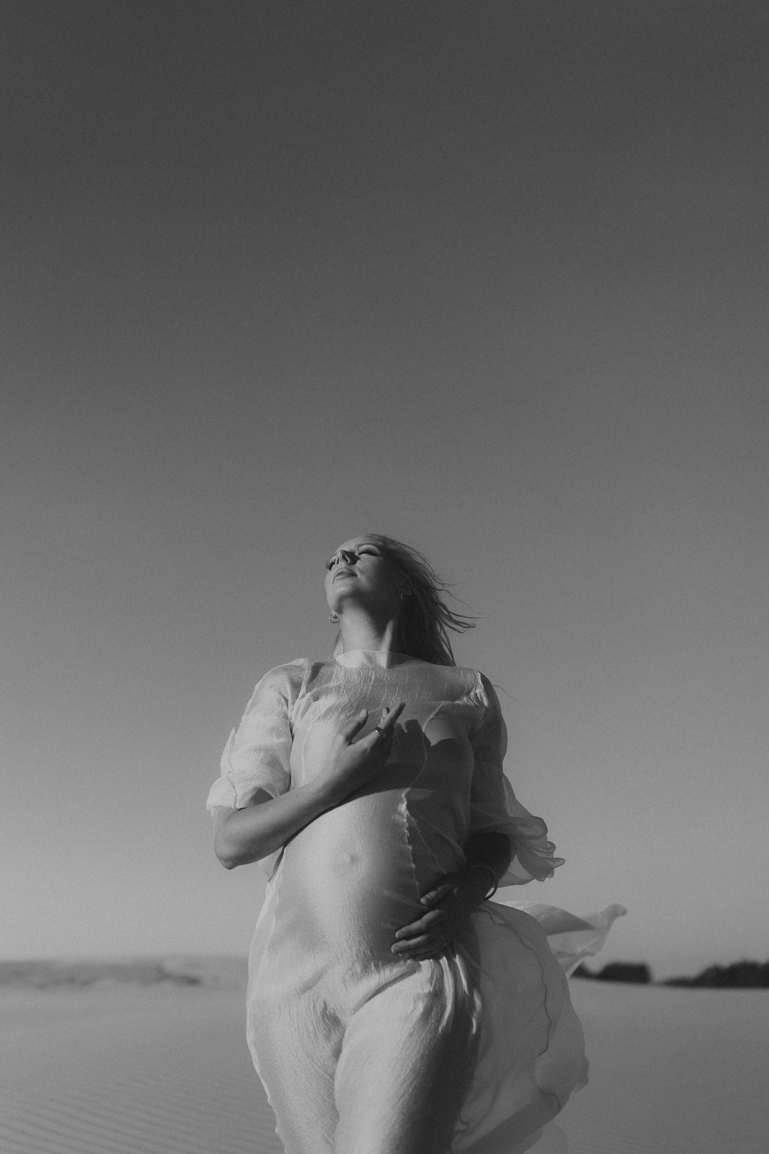 Black and white photo of a pregnant woman in flowing white dress standing in desert dunes, head tilted back.